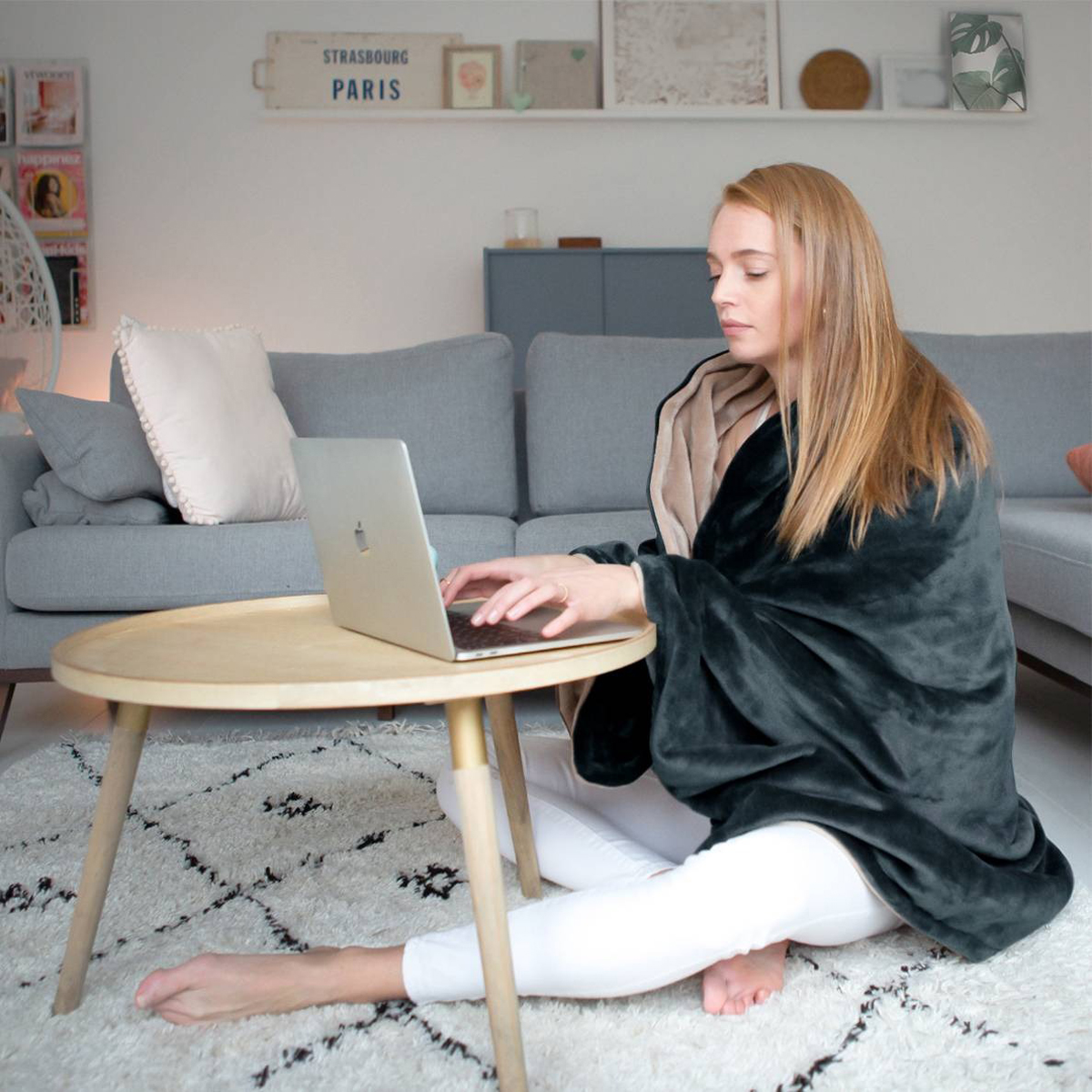 Woman sits on a rug, using a laptop, wrapped in a dark blanket.