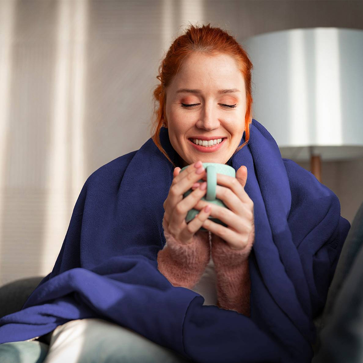 Femme enveloppée dans une couverture bleue, tenant une tasse, souriant chaleureusement, avec un cadre intérieur et un éclairage doux.
