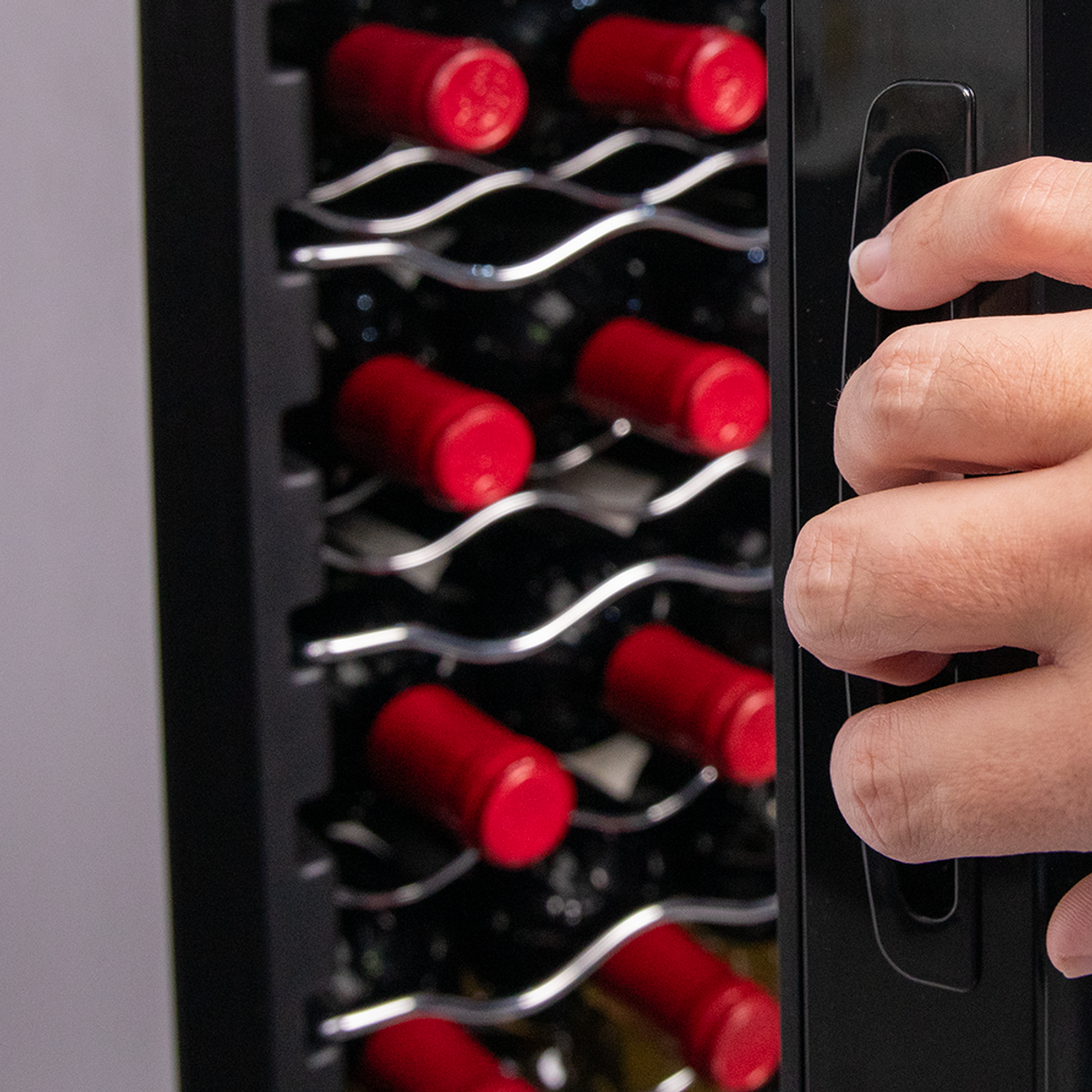 Una mano che apre la porta di un wine cooler nero, rivelando bottiglie con tappi rossi all'interno.