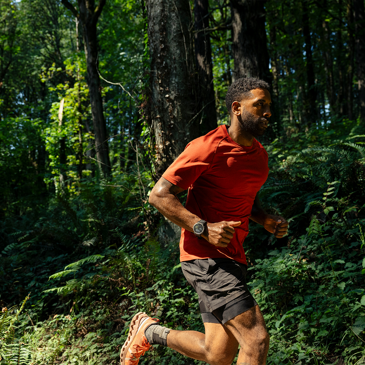 Mężczyzna biegnący w lesie. Ma na sobie czerwoną koszulę, czarne szorty i zegarek. Zielone drzewa w tle.