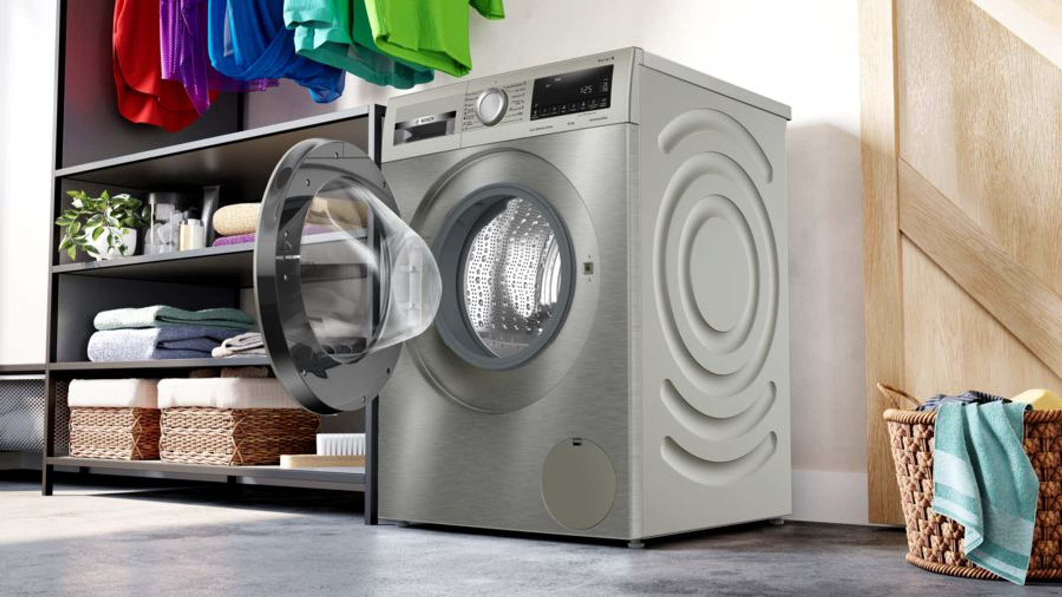 Srebrna pralka z otwartymi drzwiami, szafka z półkami i koszami na pranie, kolorowe ubrania.