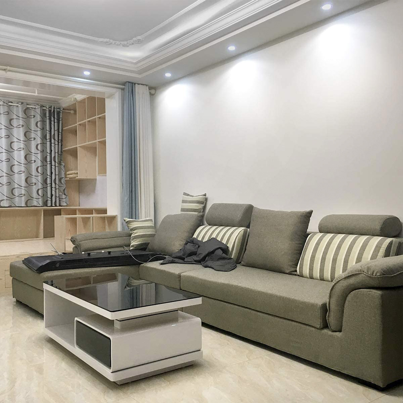 Wohnzimmer mit weißen Wänden, grauem Sofa, Couchtisch und Holzregalen mit Lichtern.