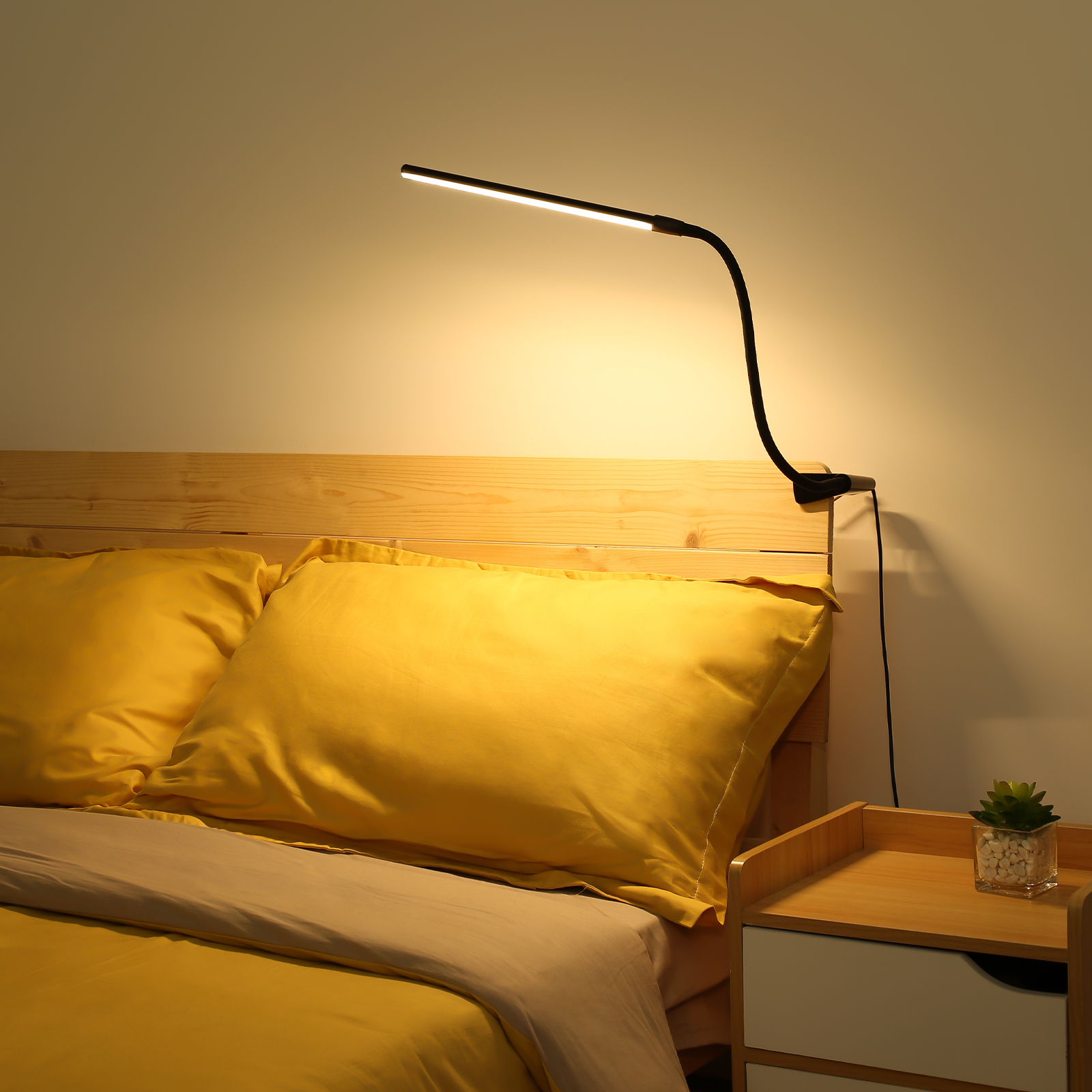 Eine schwarze, flexible LED-Lampe an einem hölzernen Bettrahmen, mit gelben Kissen und einer kleinen Pflanze.