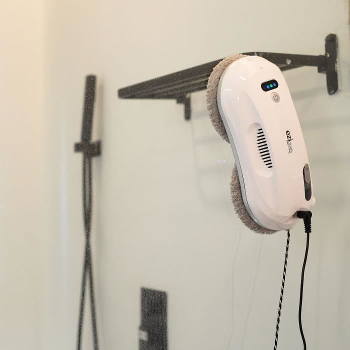 Biała myjka do okien z czarnym kablem znajduje się na szybie, a w tle czarna armatura prysznicowa.