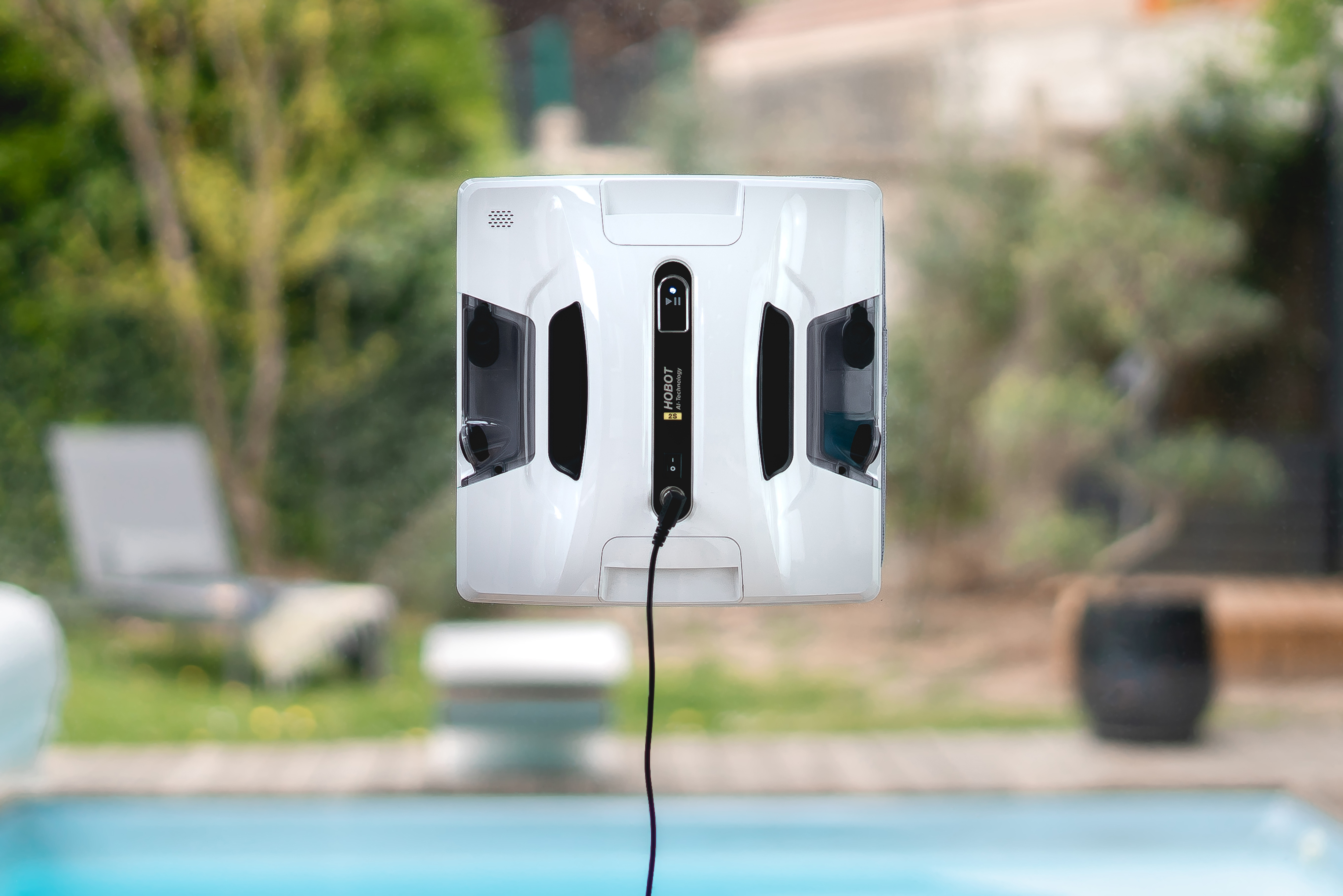 Weißer Roboter-Fensterputzer auf Glas, Blick auf Pool und Garten im unscharfen Hintergrund.