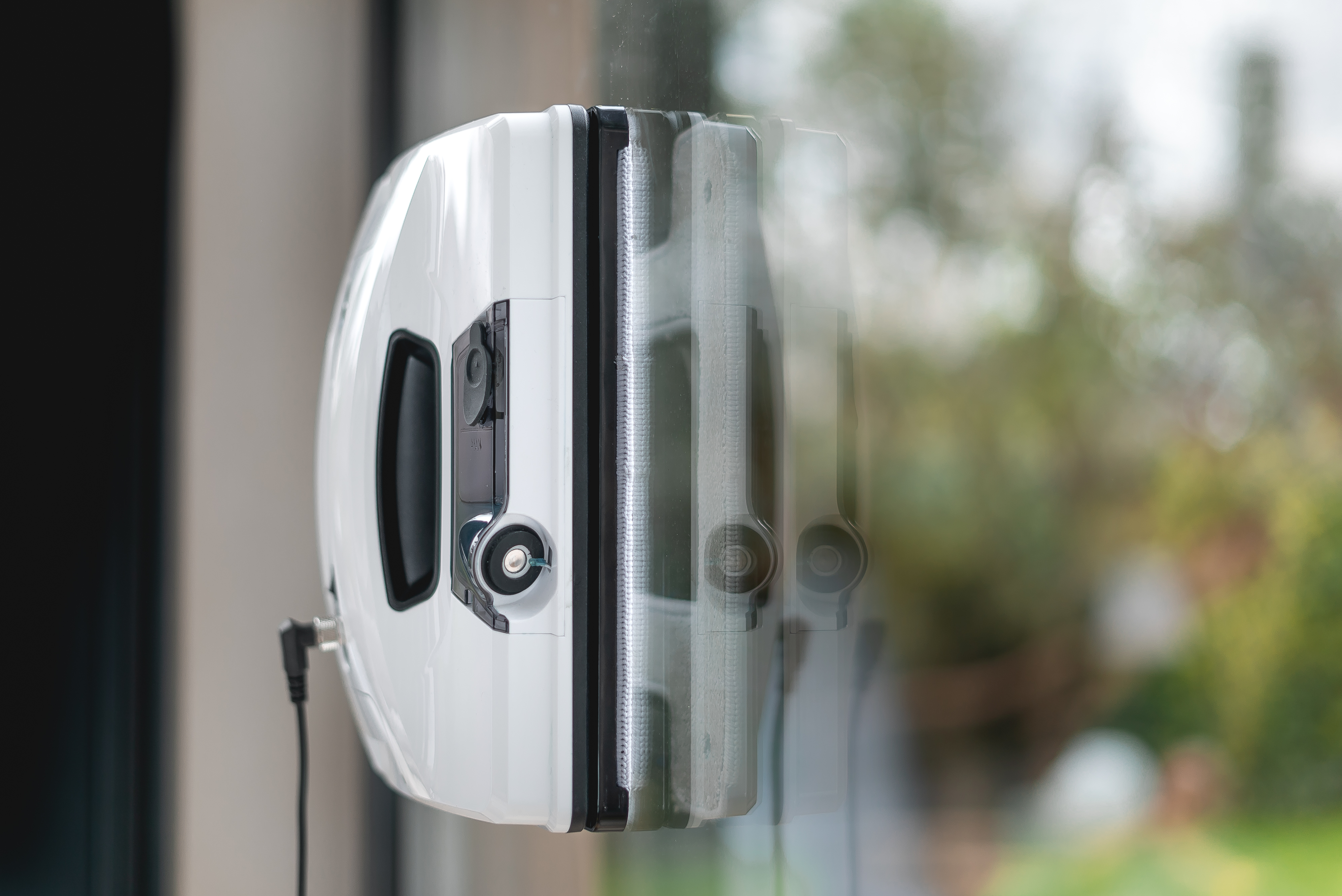 Weißer Fensterreiniger an einem Fenster befestigt. Er hat ein Kabel und reinigt das Fenster.