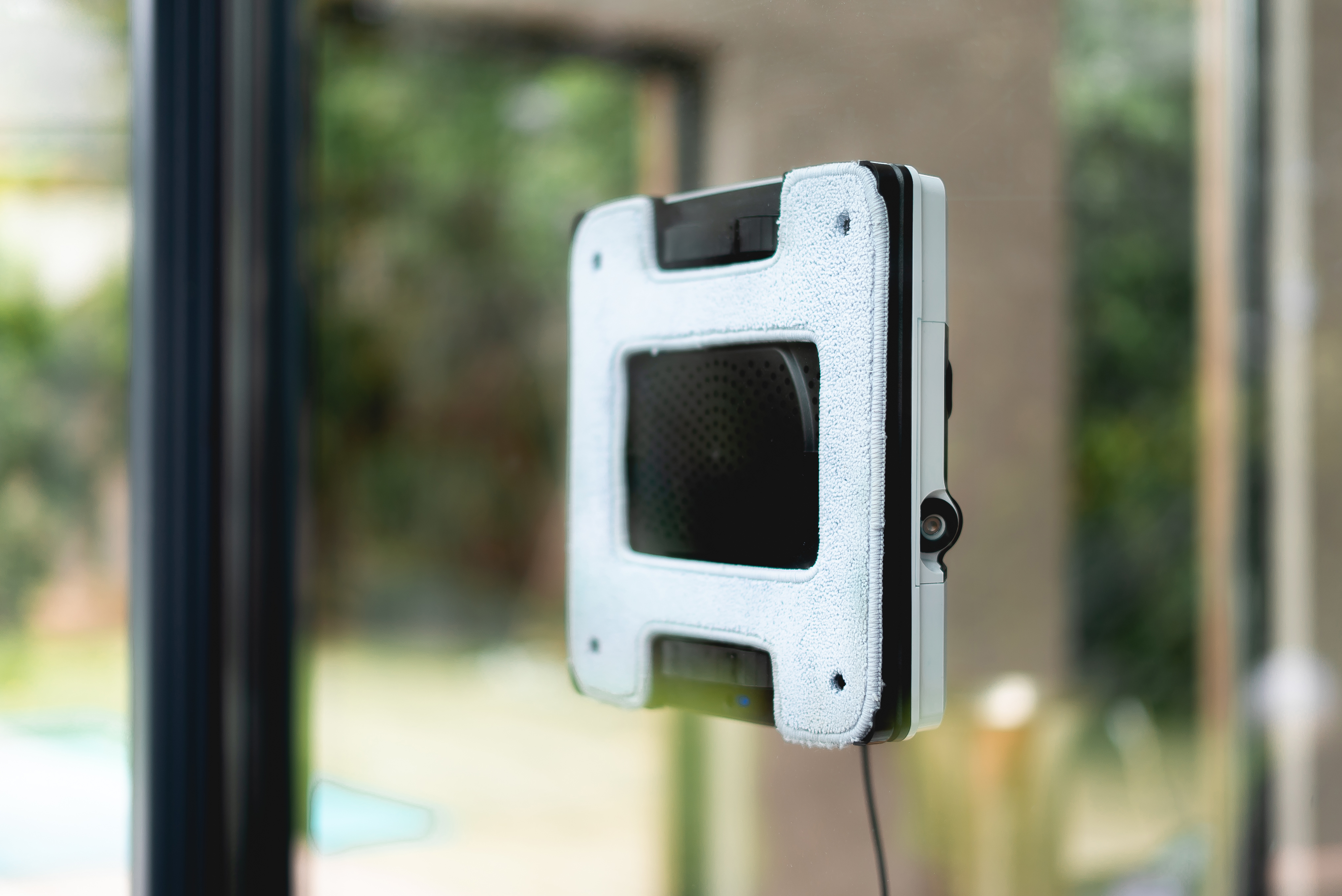 Ein weißer und schwarzer Fensterputzroboter, der an einem Fenster befestigt ist. Er reinigt das Glas.