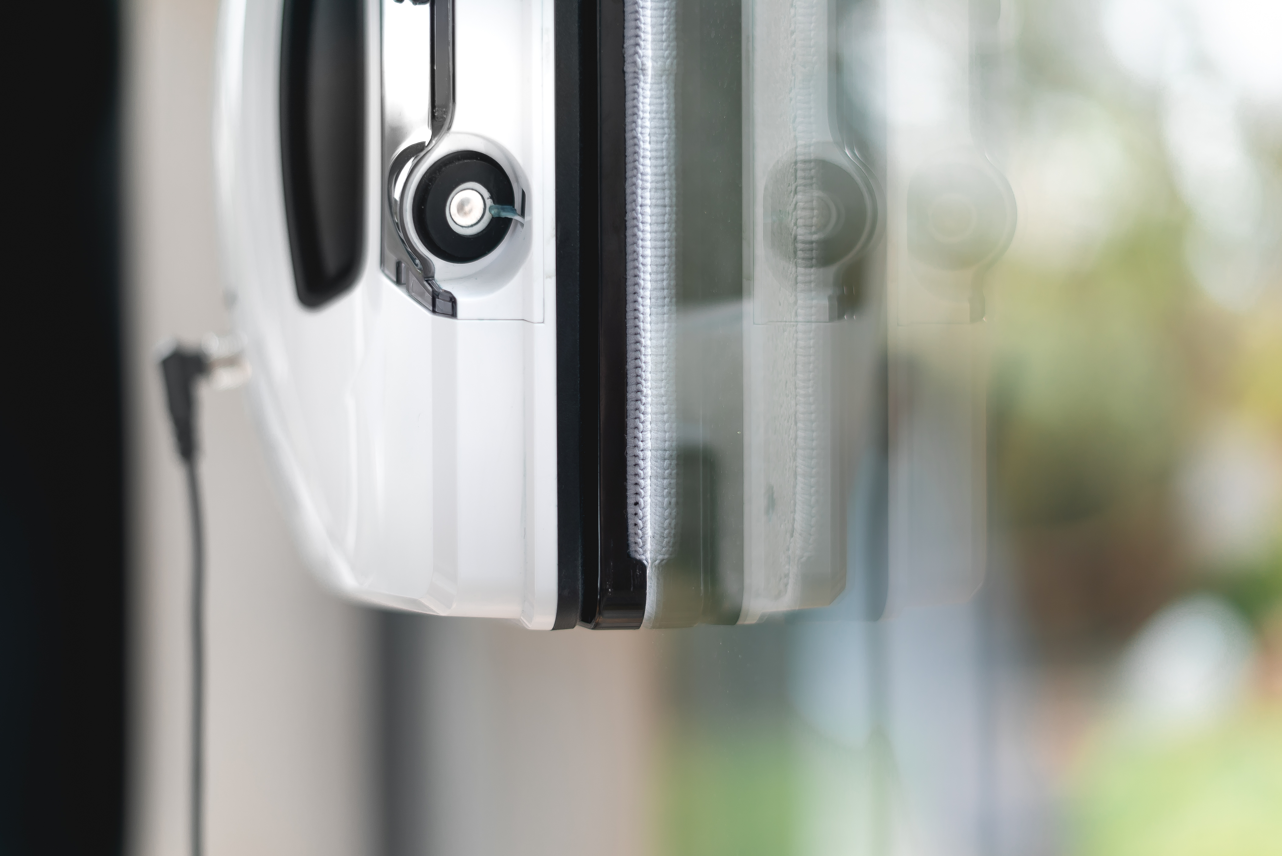 Ein weißer Fensterreiniger mit schwarzem Kabel, an einem Fenster befestigt, mit verschwommenem Hintergrund.