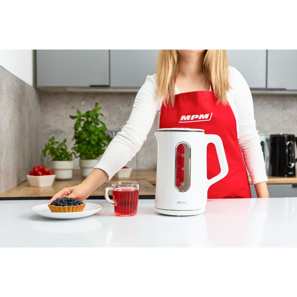 Donna con grembiule rosso, bollitore bianco, che tiene una crostata e una tazza di liquido rosso su un tavolo.