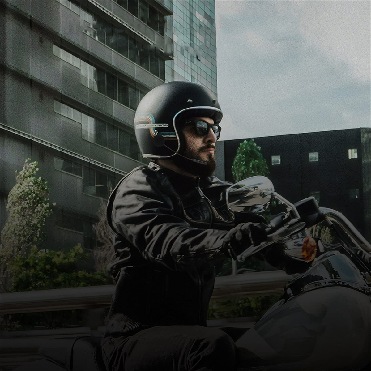 Mann auf Motorrad mit schwarzem Helm und Sonnenbrille, fährt in der Nähe eines hohen Gebäudes.