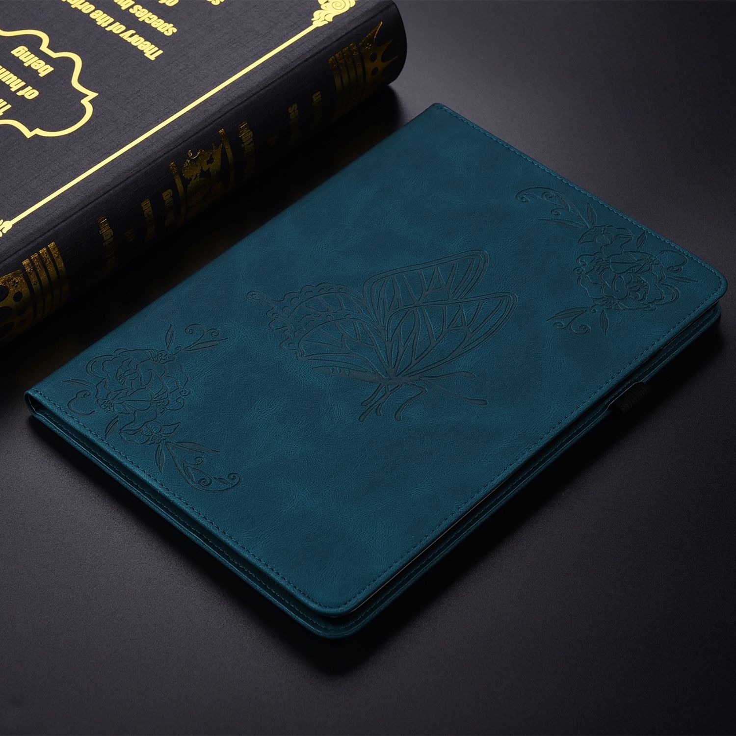 Türkisfarbene Leder-Tablethülle mit Schmetterlingsdesign und Blumenakzenten, neben einem Buch.