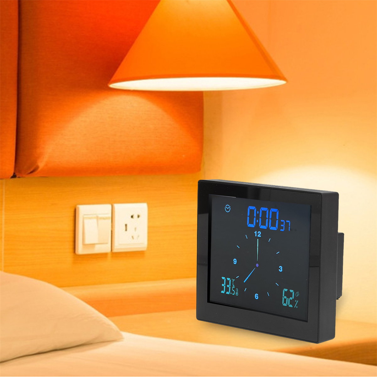 Eine schwarze Digitaluhr zeigt Zeit und Temperatur auf einem Nachttisch mit einer Lampe darüber.