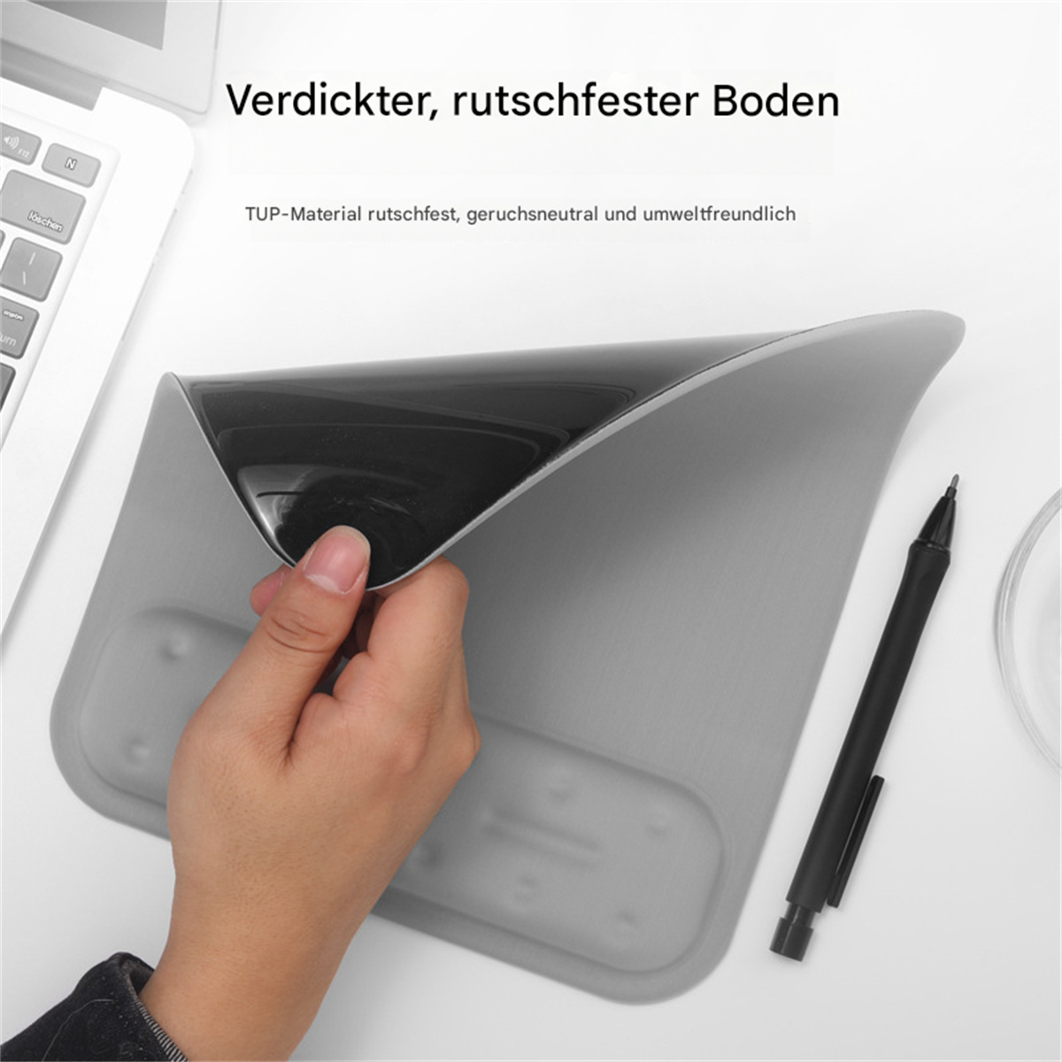 Eine Hand hält den Rand eines grauen Mauspads, Laptop und Stift in der Nähe.