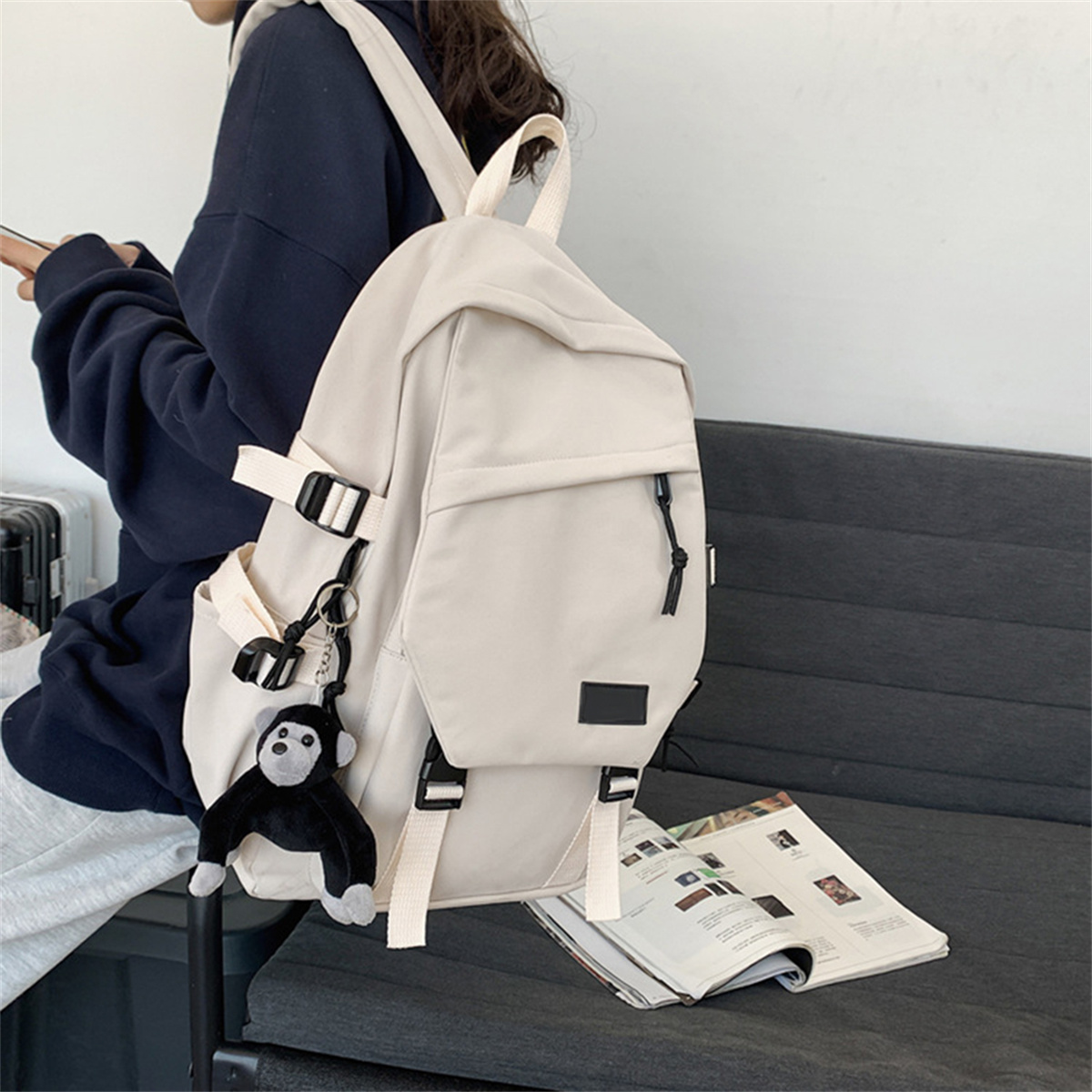 Beiger Rucksack mit schwarzen Trägern und einem Affen-Schlüsselanhänger, auf Sofa neben Magazin.