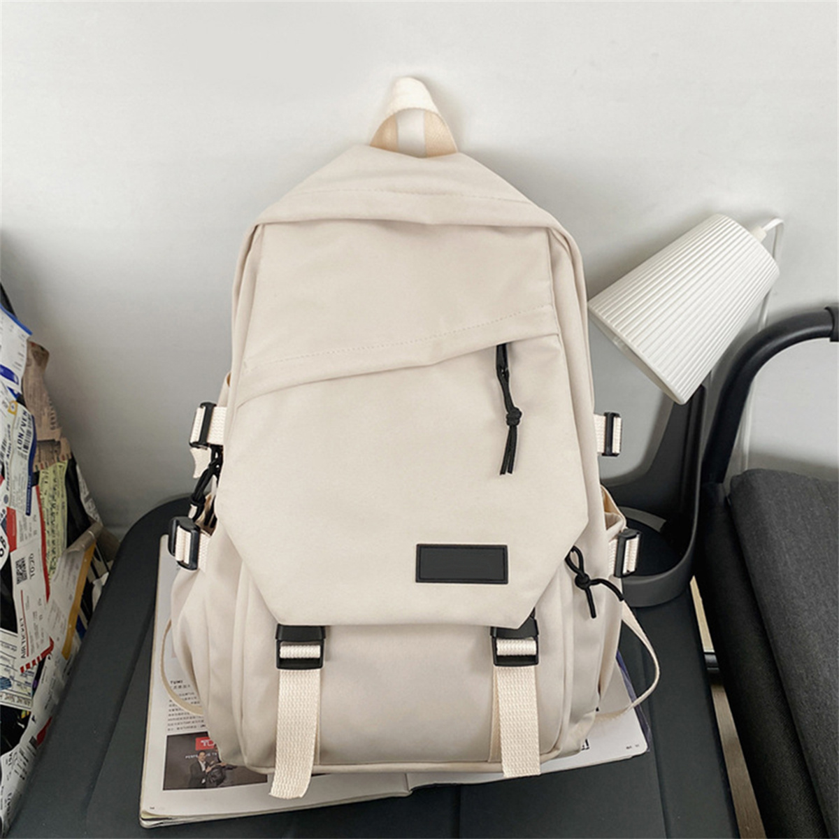 Ein cremefarbener Rucksack mit mehreren Riemen, Schnallen und einem kleinen schwarzen Etikett. Er steht an einer Wand.
