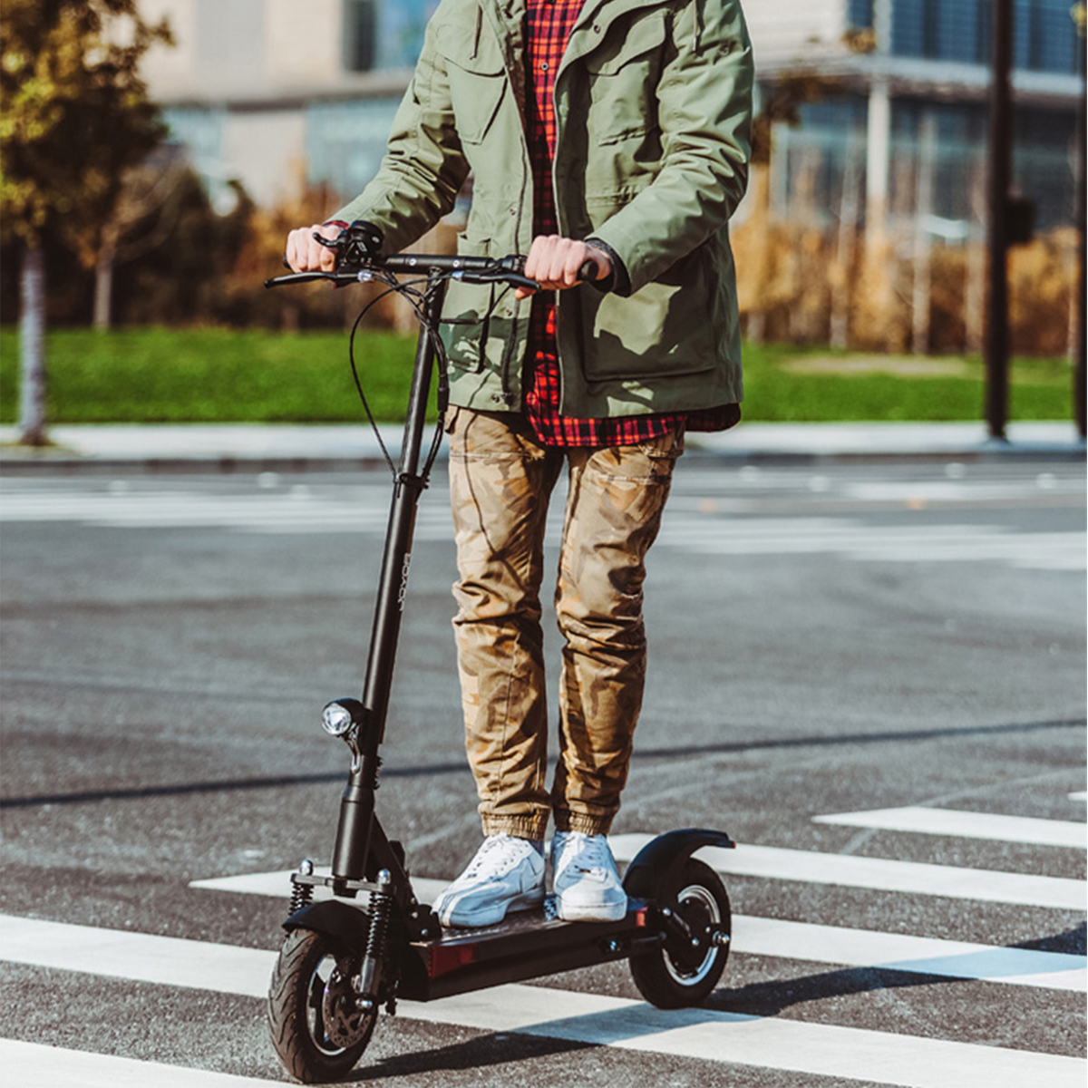 Ein Mann auf einem Elektroroller auf einem Zebrastreifen. Er trägt eine grüne Jacke und braune Hosen.