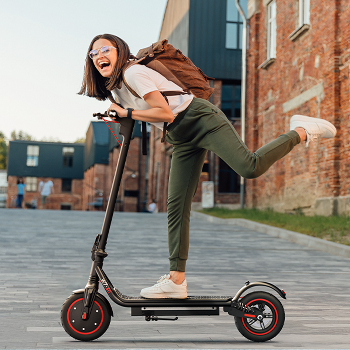 Młoda kobieta jedzie czarną hulajnogą, uśmiechając się w okularach. Jest przed ceglanym budynkiem.