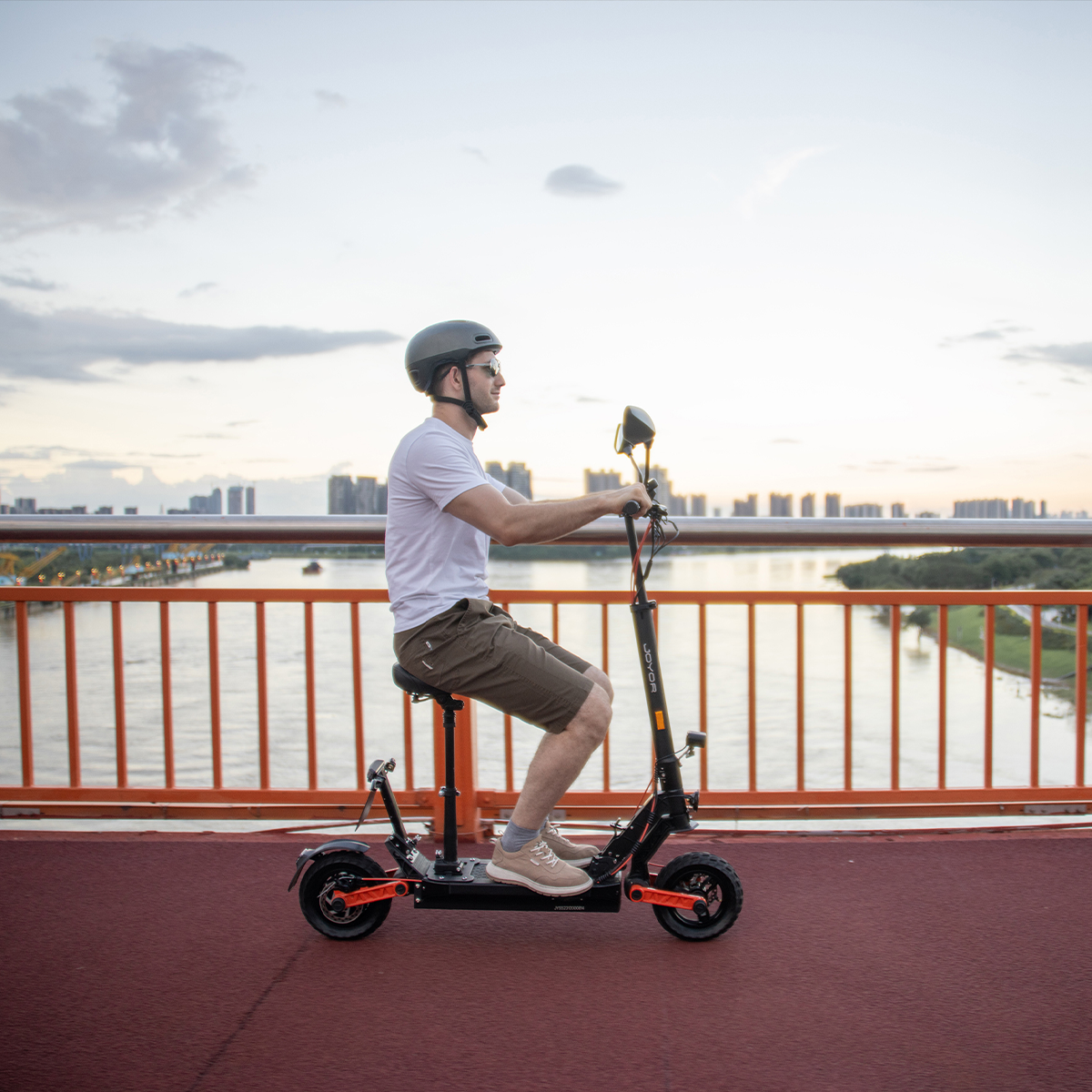 Uomo che guida uno scooter elettrico nero e arancione su un ponte con fiume ed edifici sullo sfondo.