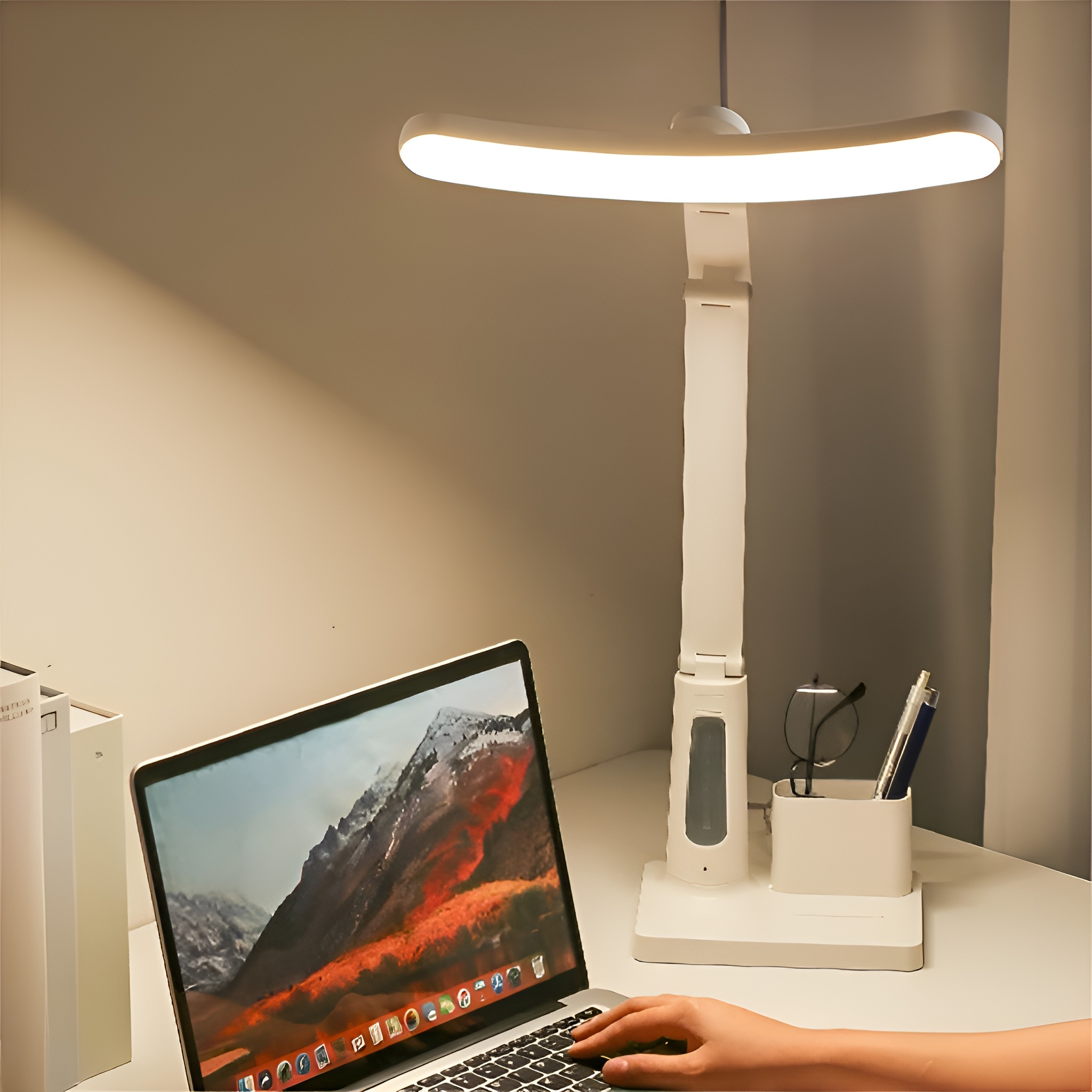 Weiße Schreibtischlampe auf dem Schreibtisch, Laptop geöffnet. Hand tippt. Brille und Stifte im Halter.