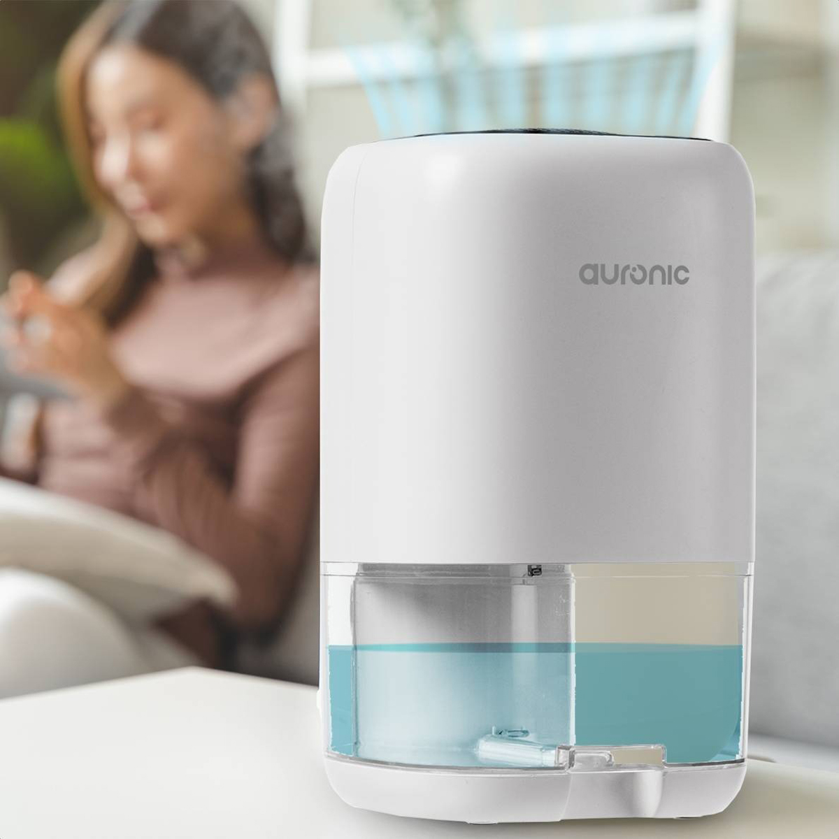 Witte luchtreiniger met zichtbare watertank, vrouw op de achtergrond gebruikt telefoon.