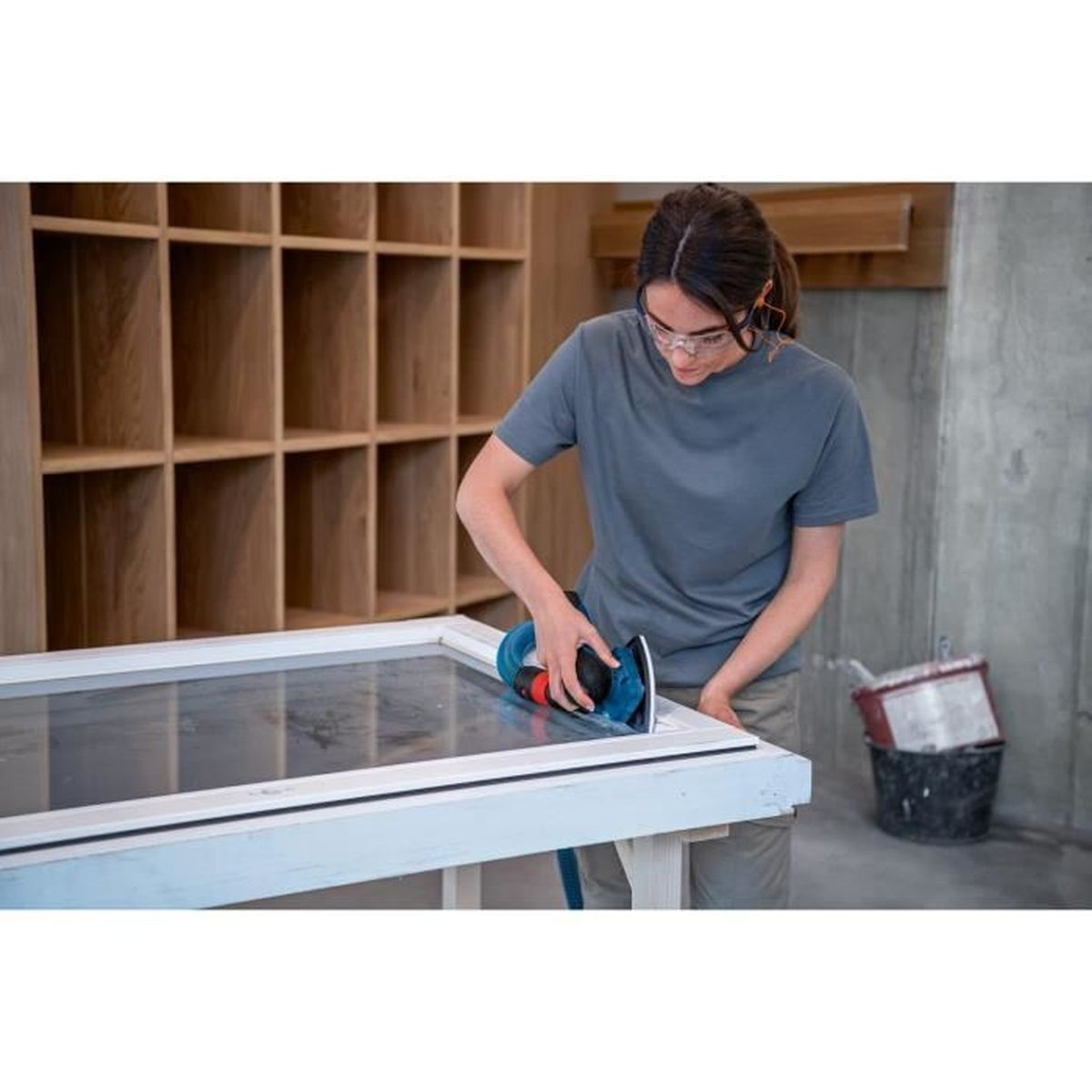 Kobieta używa szlifierki elektrycznej na białej powierzchni. Ma na sobie okulary ochronne.