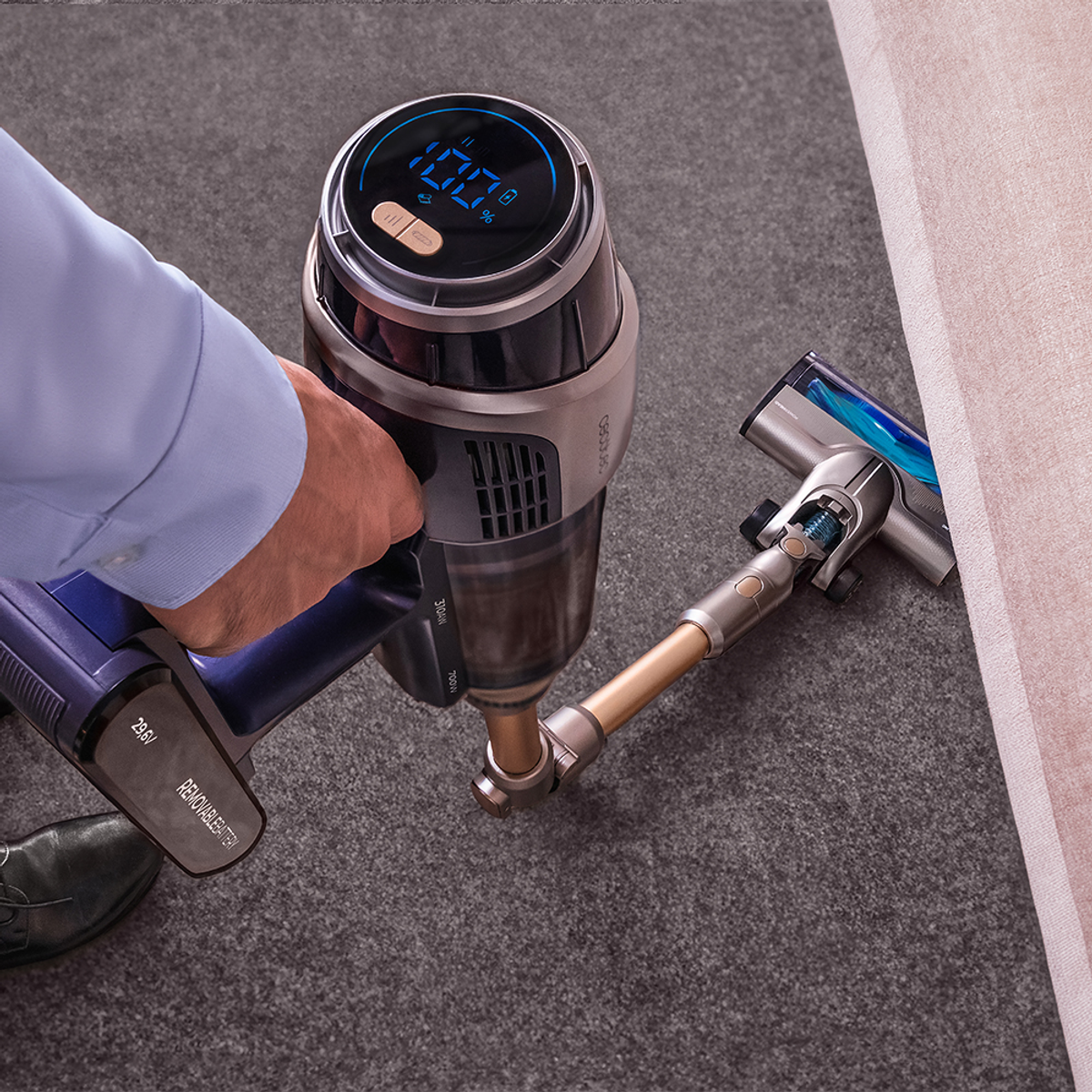 Una persona usando una aspiradora de mano azul y plateada en una alfombra gris cerca de un sofá beige.