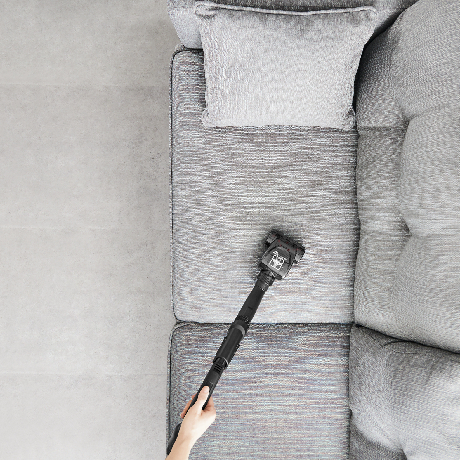 Ręka odkurza szarą sofę. Głowica odkurzacza jest blisko powierzchni sofy.