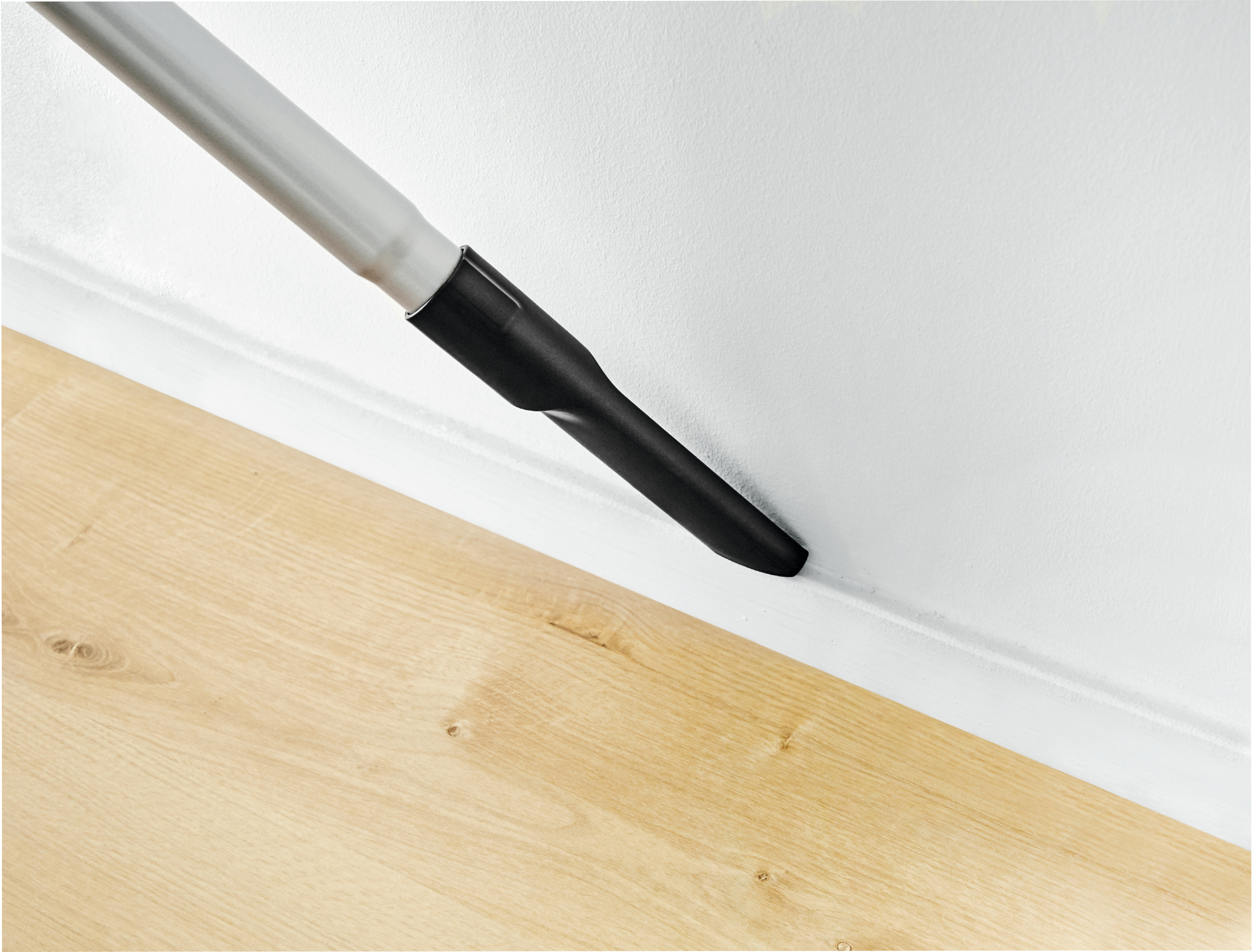 Dysza odkurzacza czyszcząca róg białej ściany, pokazująca drewnianą podłogę i białą listwę.