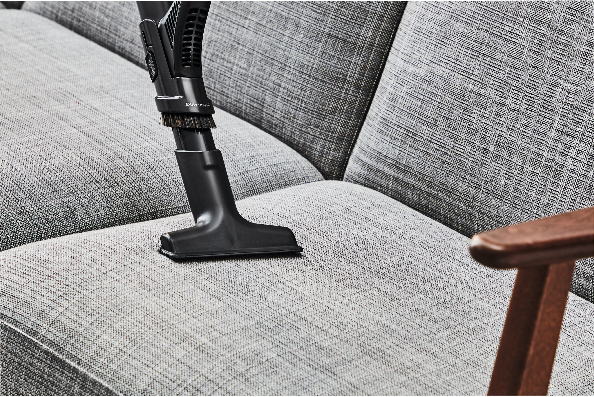 Odkurzacz czyszczący szarą sofę z drewnianym krzesłem w rogu.