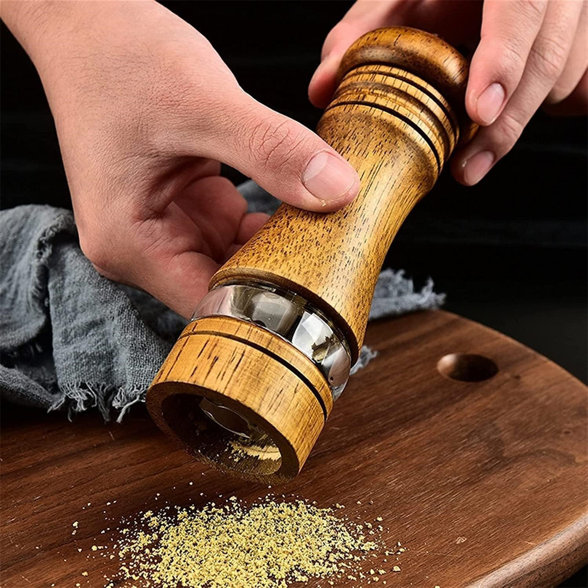 Hände halten eine hölzerne Pfeffermühle über einem hölzernen Schneidebrett mit Pfefferkörnern.