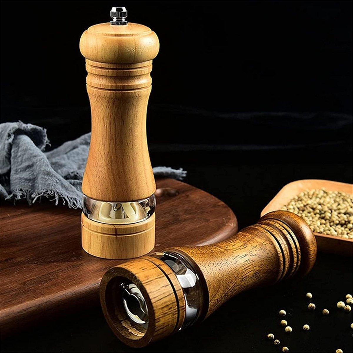 Zwei hölzerne Pfeffermühlen mit Glasteil, auf einem Holzbrett mit Pfefferkörnern und dunklem Hintergrund.