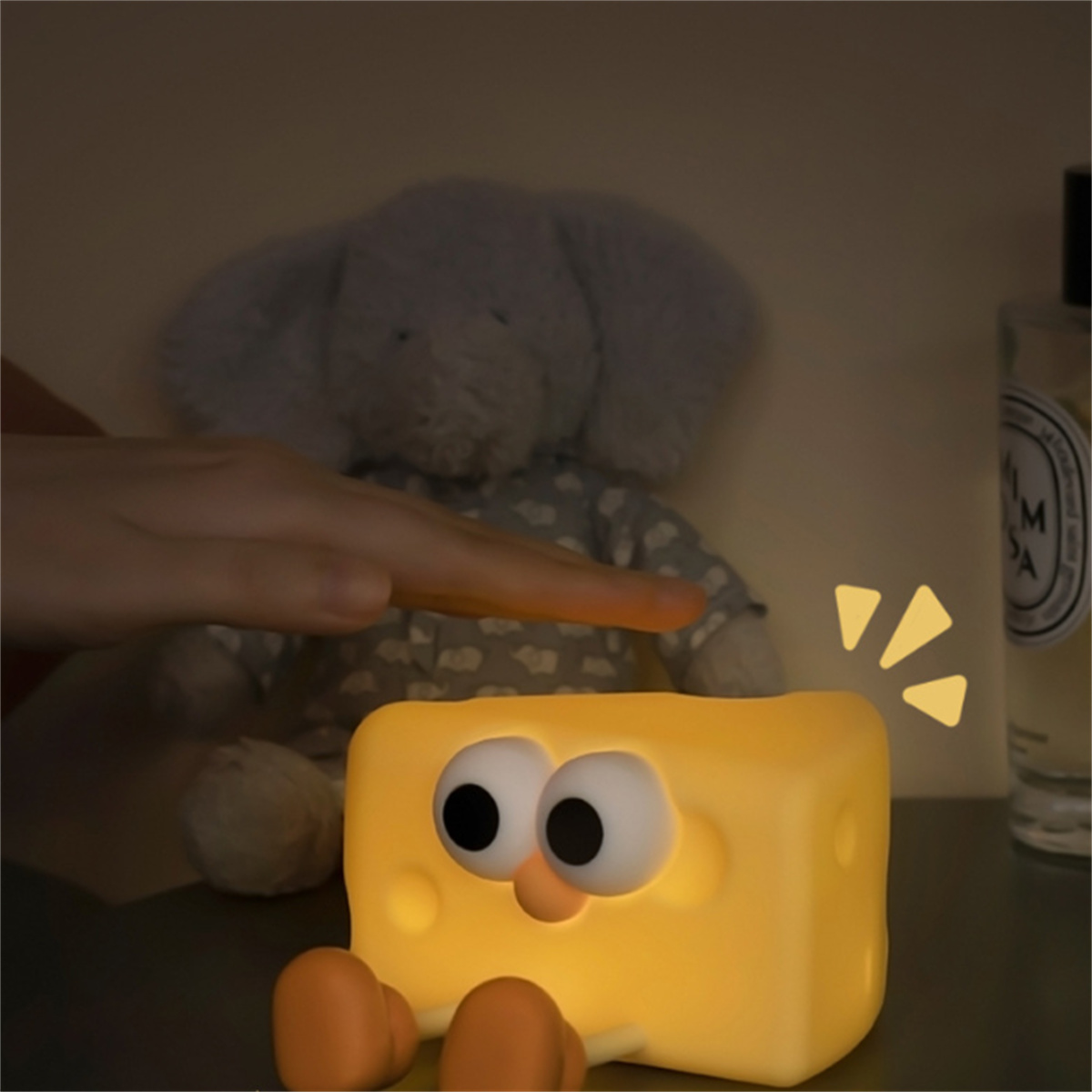 Eine käseförmige Lampe mit Kulleraugen, eine Hand darüber und ein Stoffelefant im Hintergrund.
