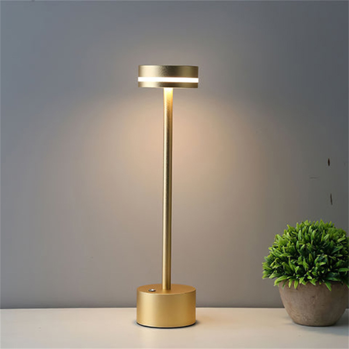 Goldfarbene Lampe. Hoher, schlanker Körper mit rundem Sockel und beleuchteter Oberseite.