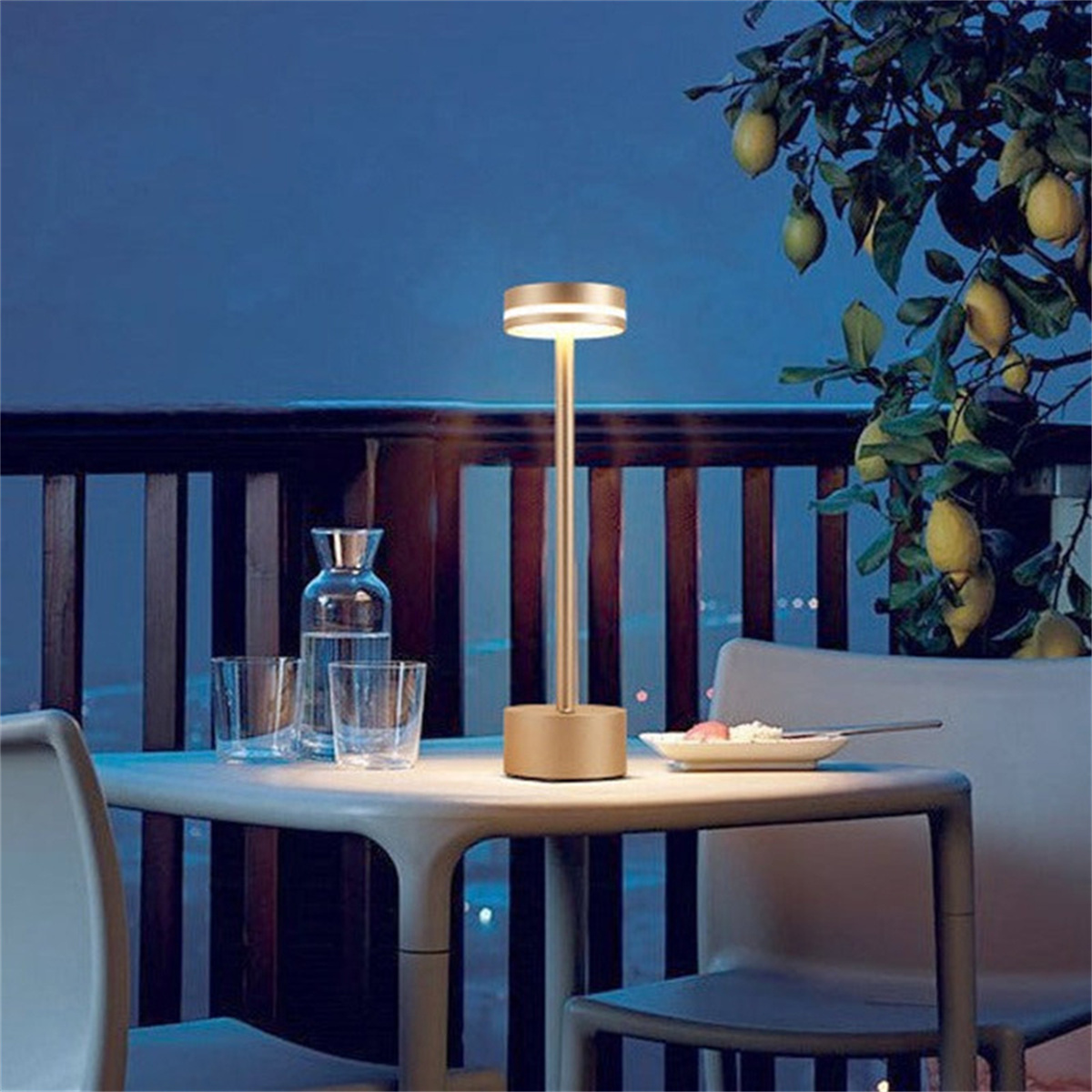 Eine Tischlampe mit Goldfinish, die einen Tisch mit Gläsern, Tellern und Zitronenbäumen im Hintergrund beleuchtet.
