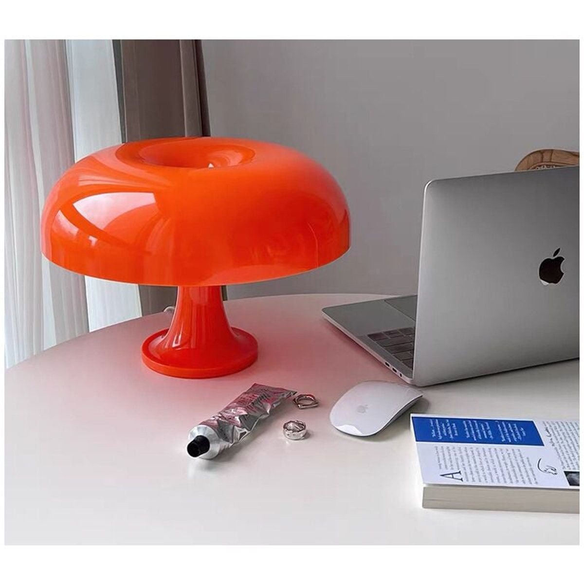 Orangefarbene pilzförmige Lampe auf einem weißen Tisch. Ein Laptop, eine Maus und eine Tube Creme sind in der Nähe.