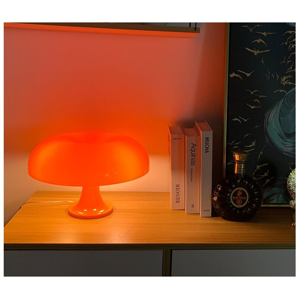 Eine orangefarbene pilzförmige Lampe auf einer Holzoberfläche, mit Büchern und einer Flasche im Hintergrund.
