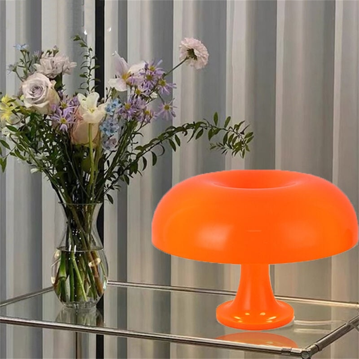 Eine orangefarbene pilzförmige Lampe neben einer Blumenvase auf einem Glastisch vor einem Vorhang.
