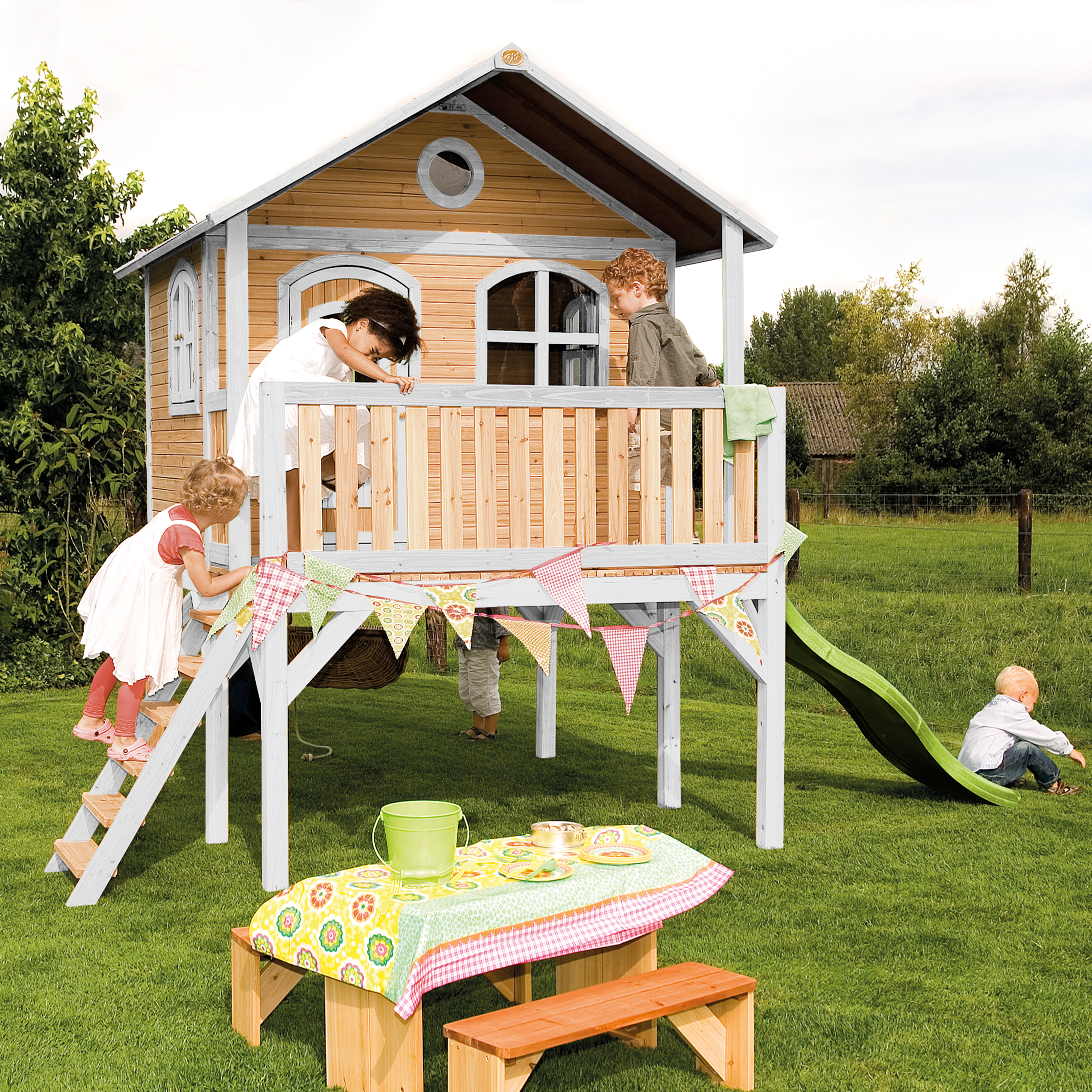 Kinder spielen auf einem Holzspielhaus mit Rutsche und Schaukel in einem grasbewachsenen Garten.