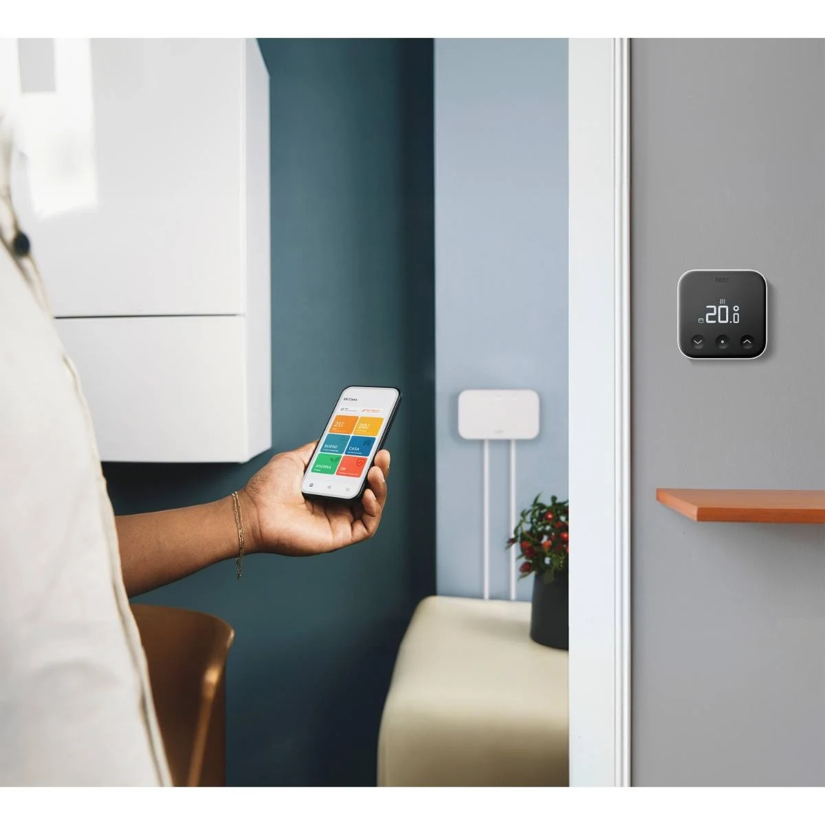 Une personne tenant un smartphone à côté d'un thermostat de maison intelligente sur un mur. La pièce a un mur bleu.