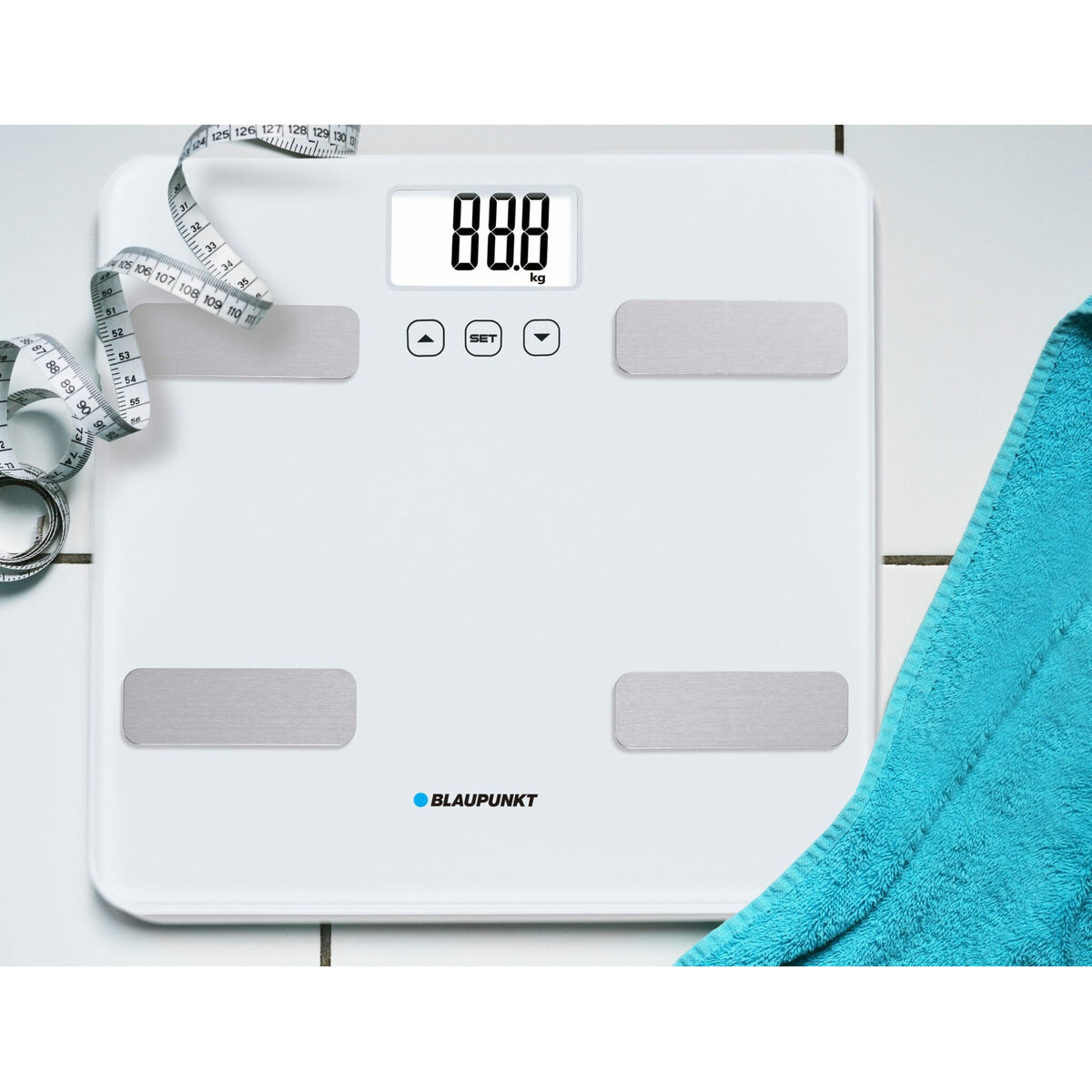 Witte weegschaal met een digitaal display en een meetlint. Een blauwe handdoek is ook in beeld.