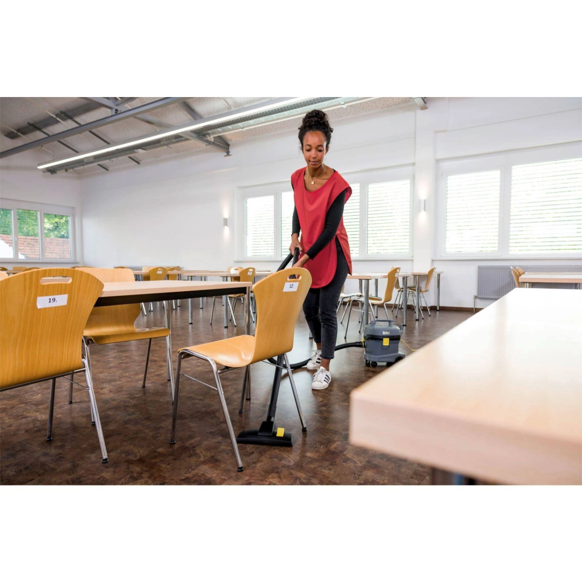 Kobieta w czerwonym sprząta podłogę w szkole odkurzaczem. Widoczne stoły i krzesła.