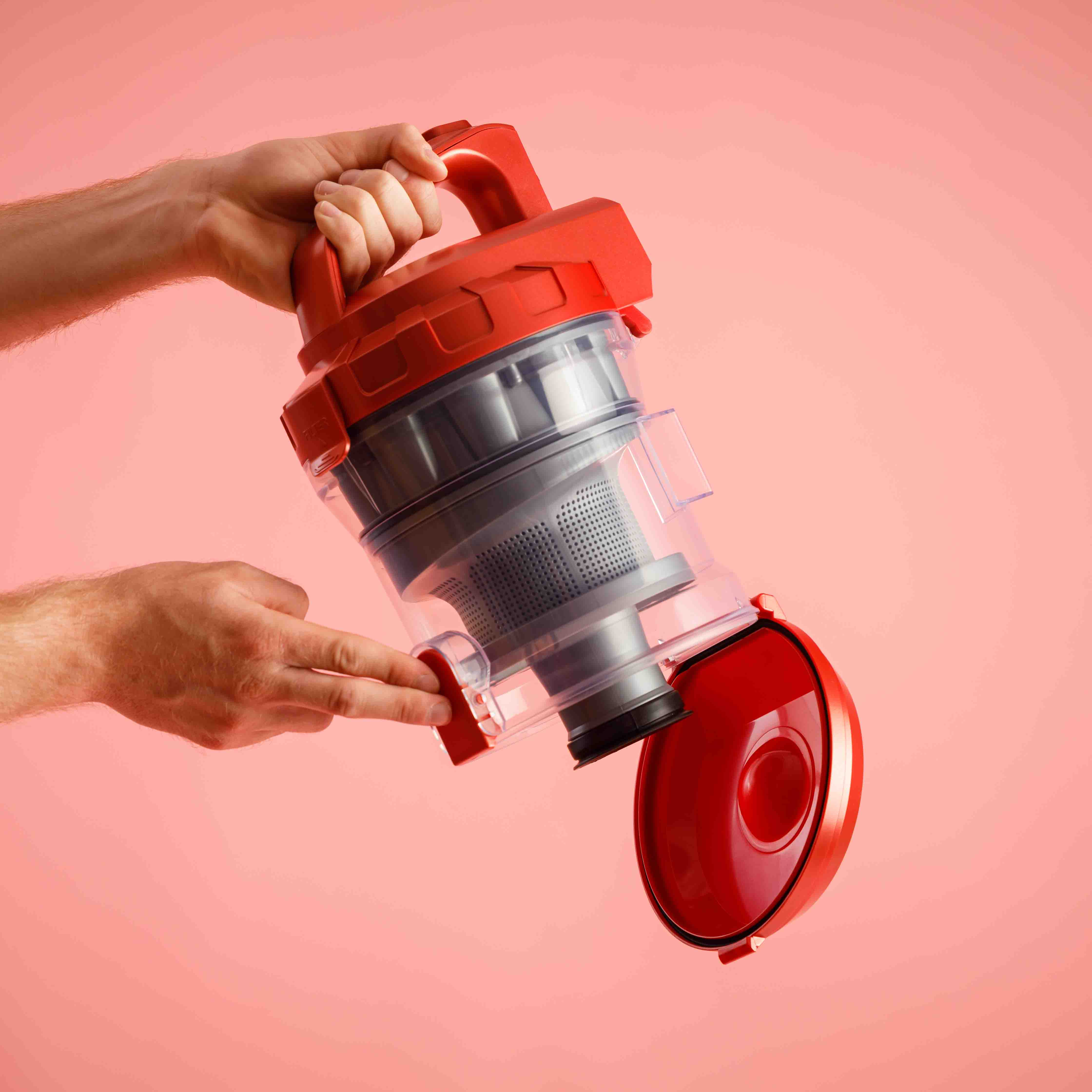 Ręce trzymające czerwony element odkurzacza, odsłaniające pojemnik na kurz. Różowe tło.