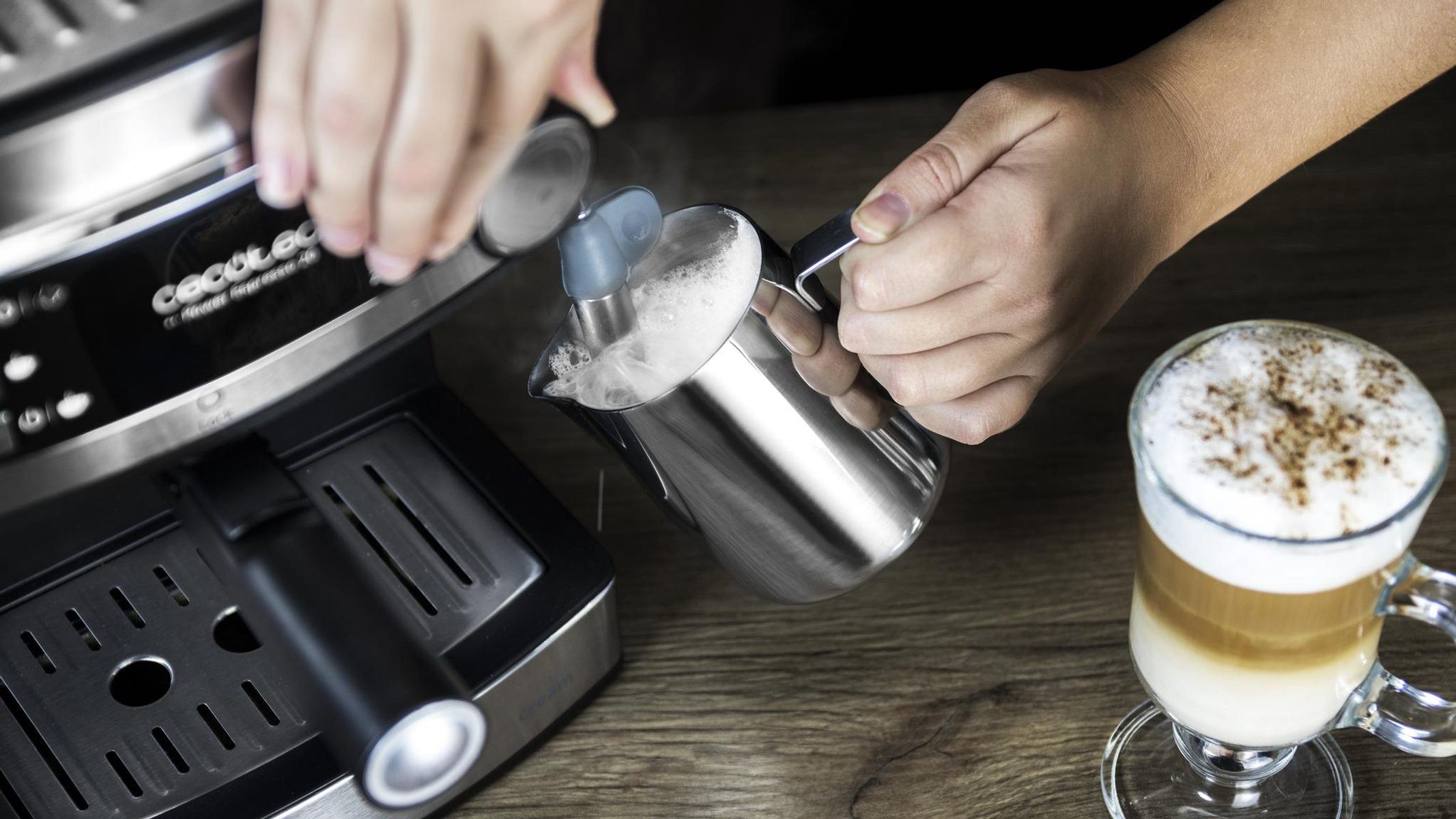 Mani che versano latte montato in una caraffa d'acciaio, vicino a una macchina per caffè espresso e un bicchiere di latte.