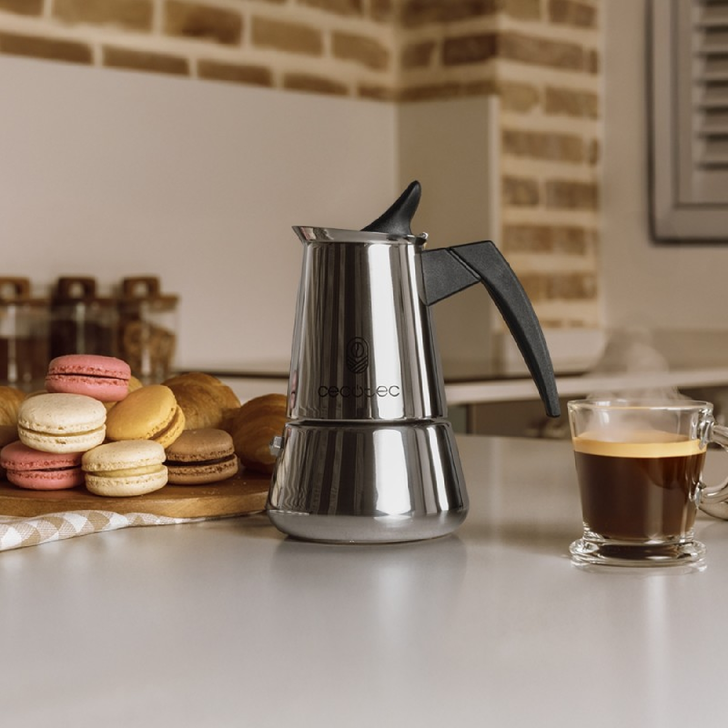 Silberner Espressokocher mit schwarzem Griff, ein Glas Kaffee und Gebäck auf weißer Oberfläche.