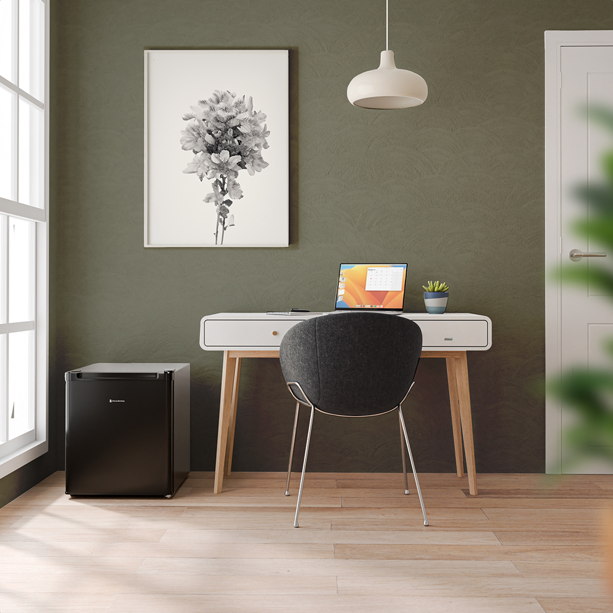 A black refrigerator next to a desk with a laptop, picture, and a plant.