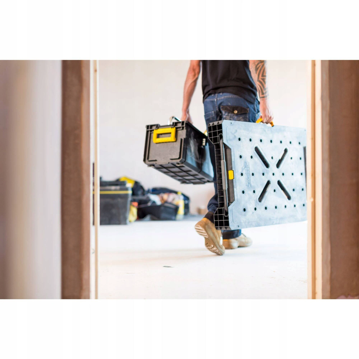 Uomo in jeans porta due cassette nere per attrezzi e un tavolo da lavoro grigio attraverso una porta.