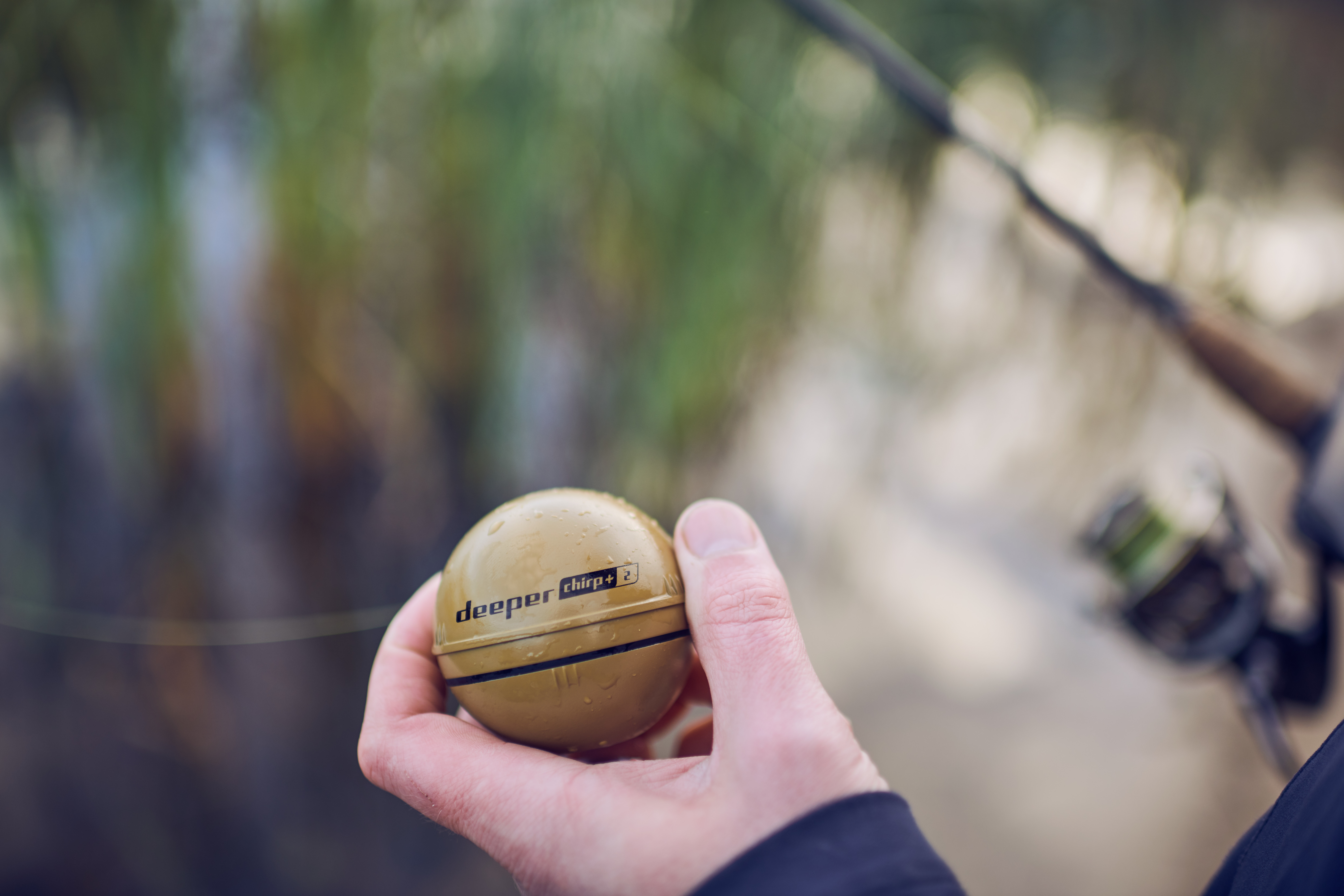 Eine Hand hält ein gelbbraunes Sonargerät über Wasser. Angelrute im Hintergrund.