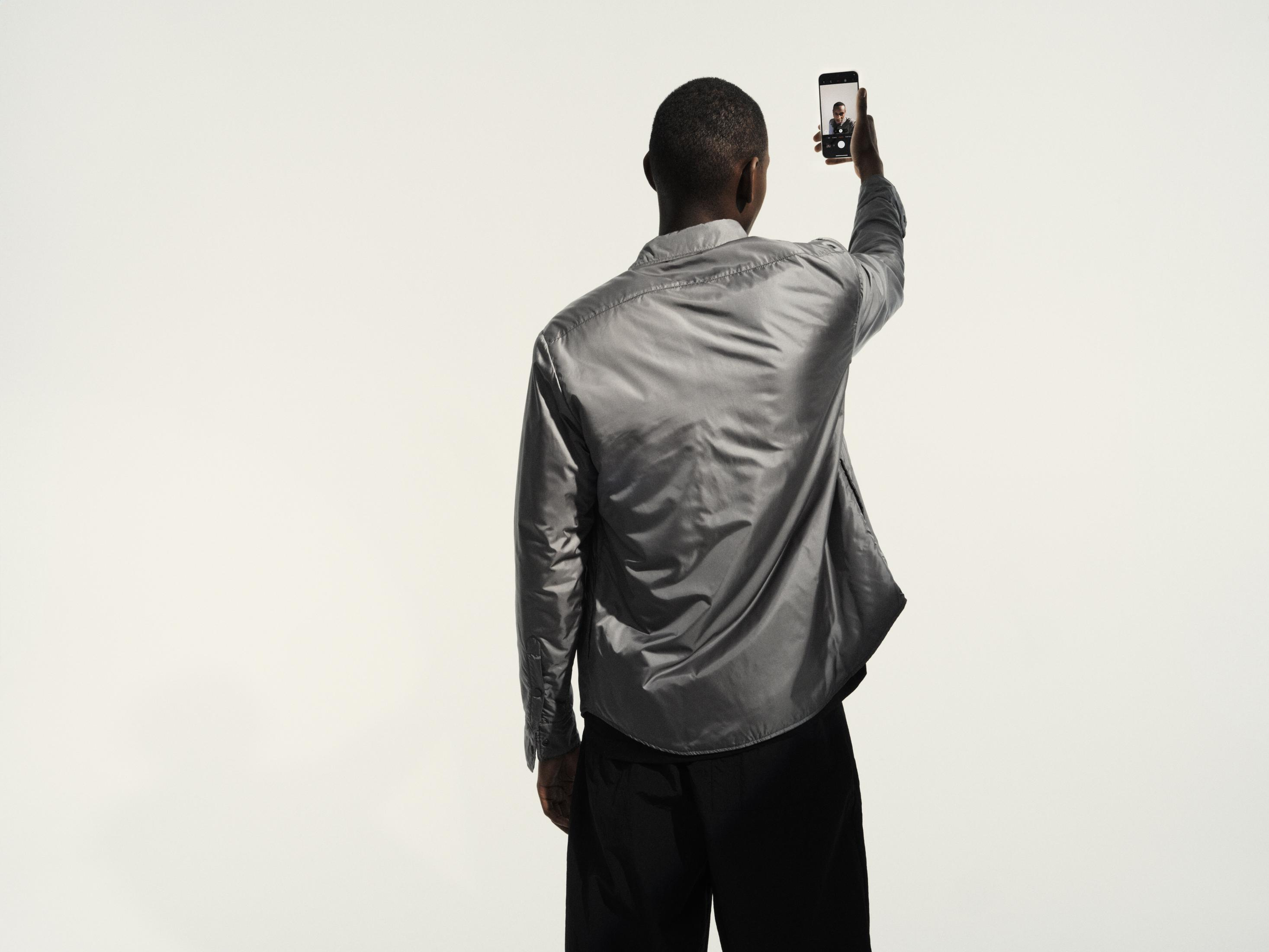 Person hält Telefon hoch, macht Selfie. Rückansicht, neutraler Hintergrund.
