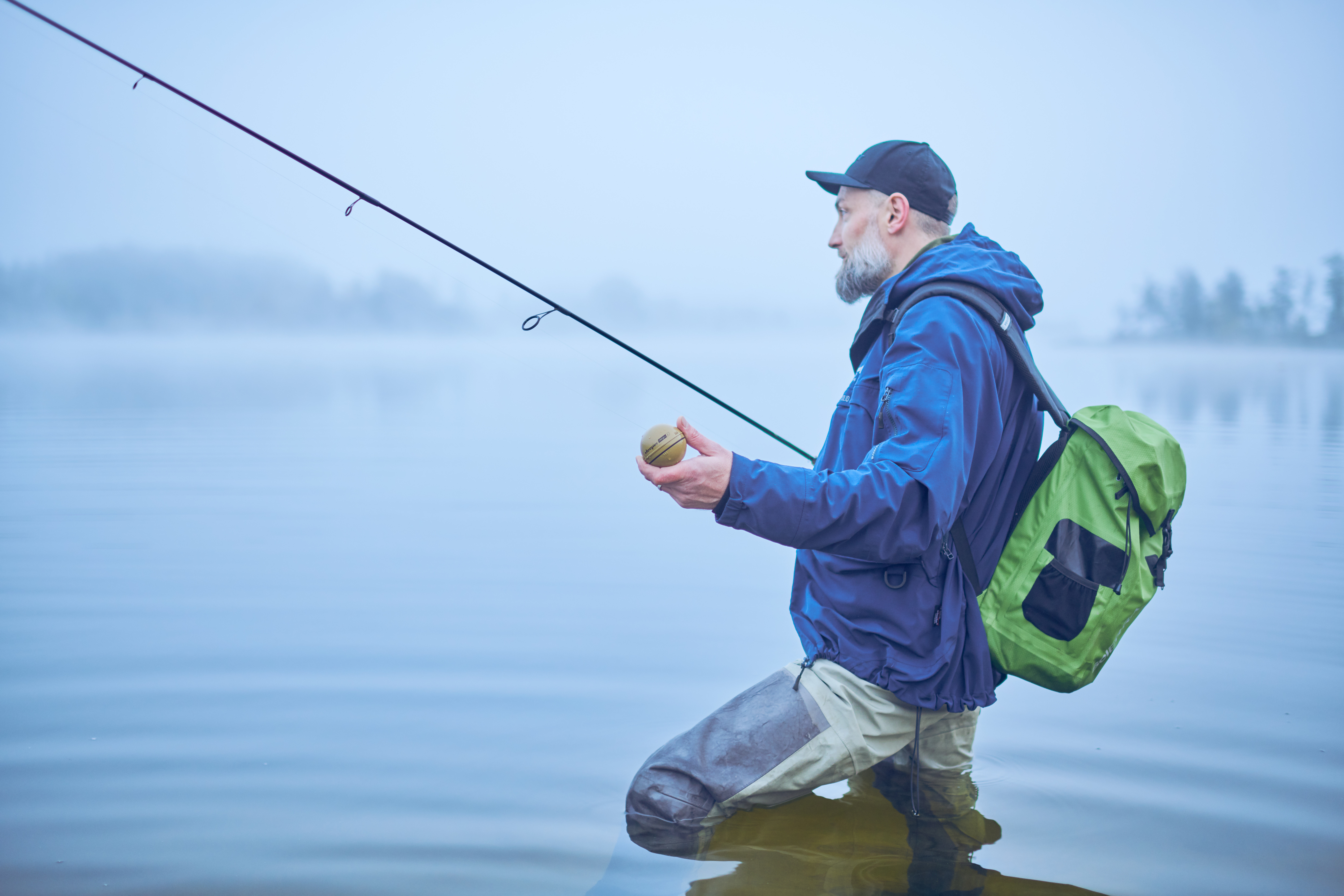 Ein Mann in einer blauen Jacke angelt in einem See. Er hält einen Köder, umgeben von Nebel.