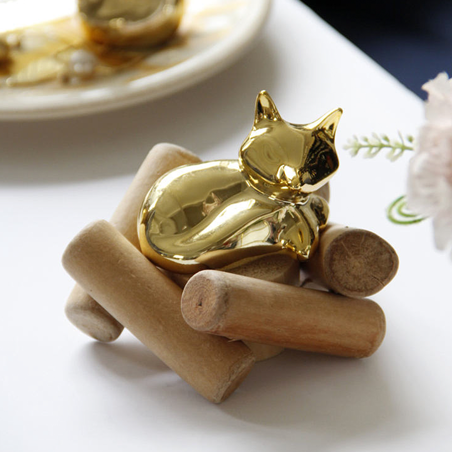 Eine goldene Katzen-Skulptur auf Holzstäben, auf weißer Oberfläche, mit unscharfem Hintergrund.